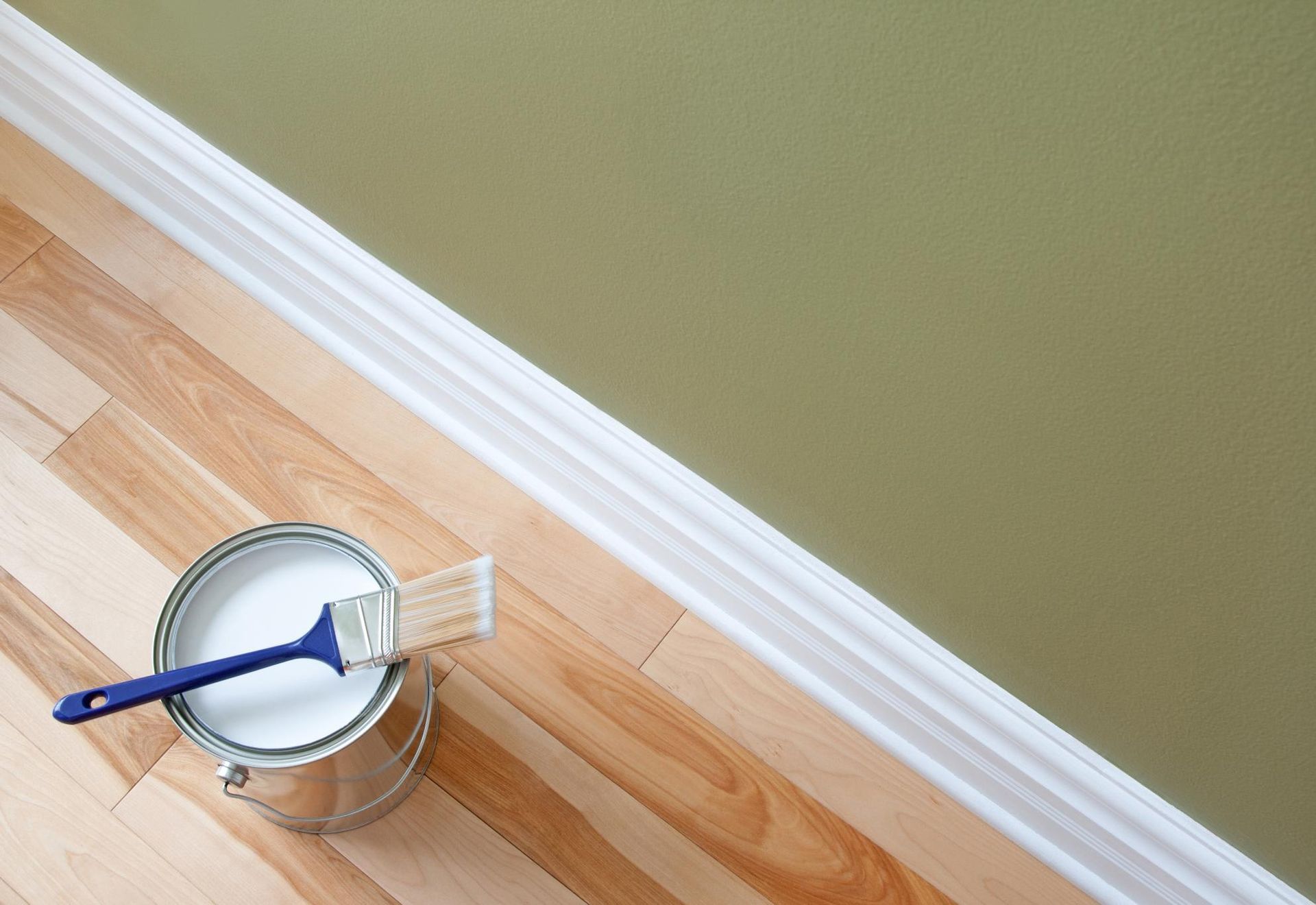 Paint can with brush on hardwood floor next to olive green wall and white trim.
