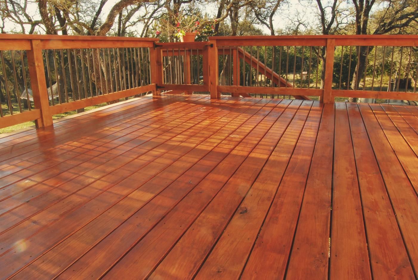 Wooden deck with reddish-brown boards and railing; the deck is in an outdoor setting with trees in the background.