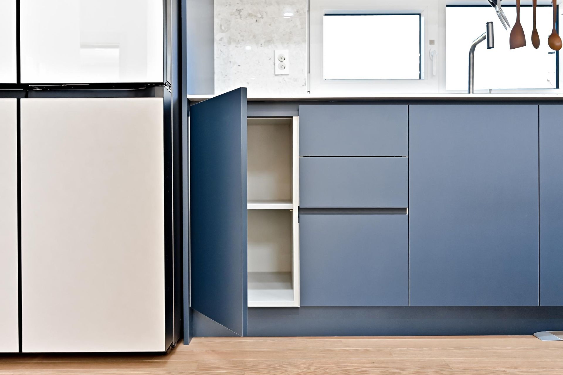 A blue kitchen cabinet is open, revealing shelves. Beside it is a cream-colored refrigerator. 
