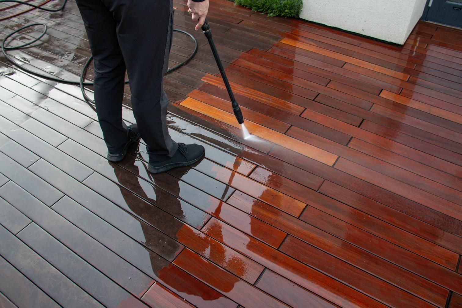 Person pressure washing a dark wooden deck, removing dirt and revealing the clean, reddish-brown wood.
