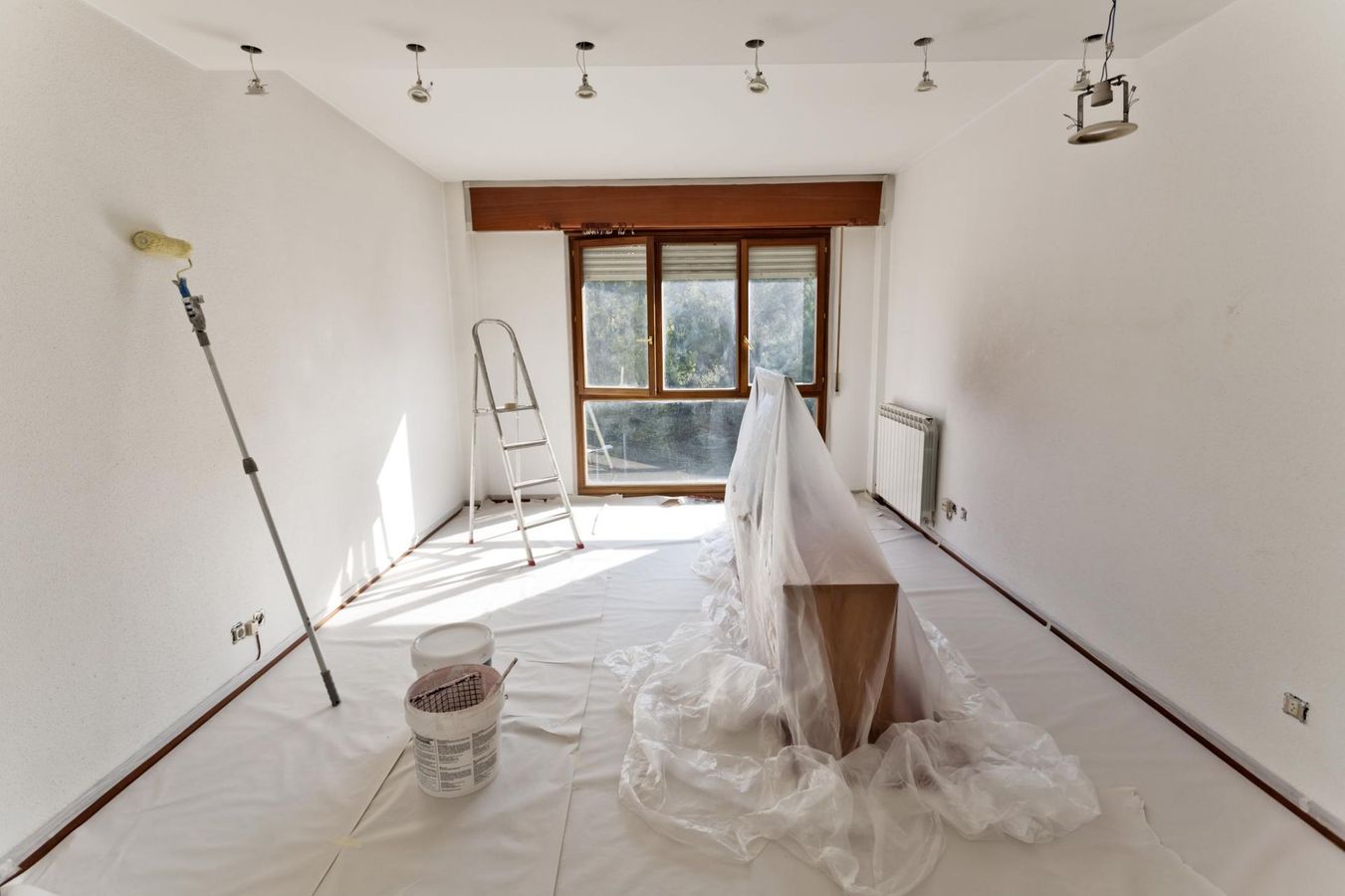 Room interior during renovation, with white walls, paint supplies, ladder, and furniture covered in plastic.