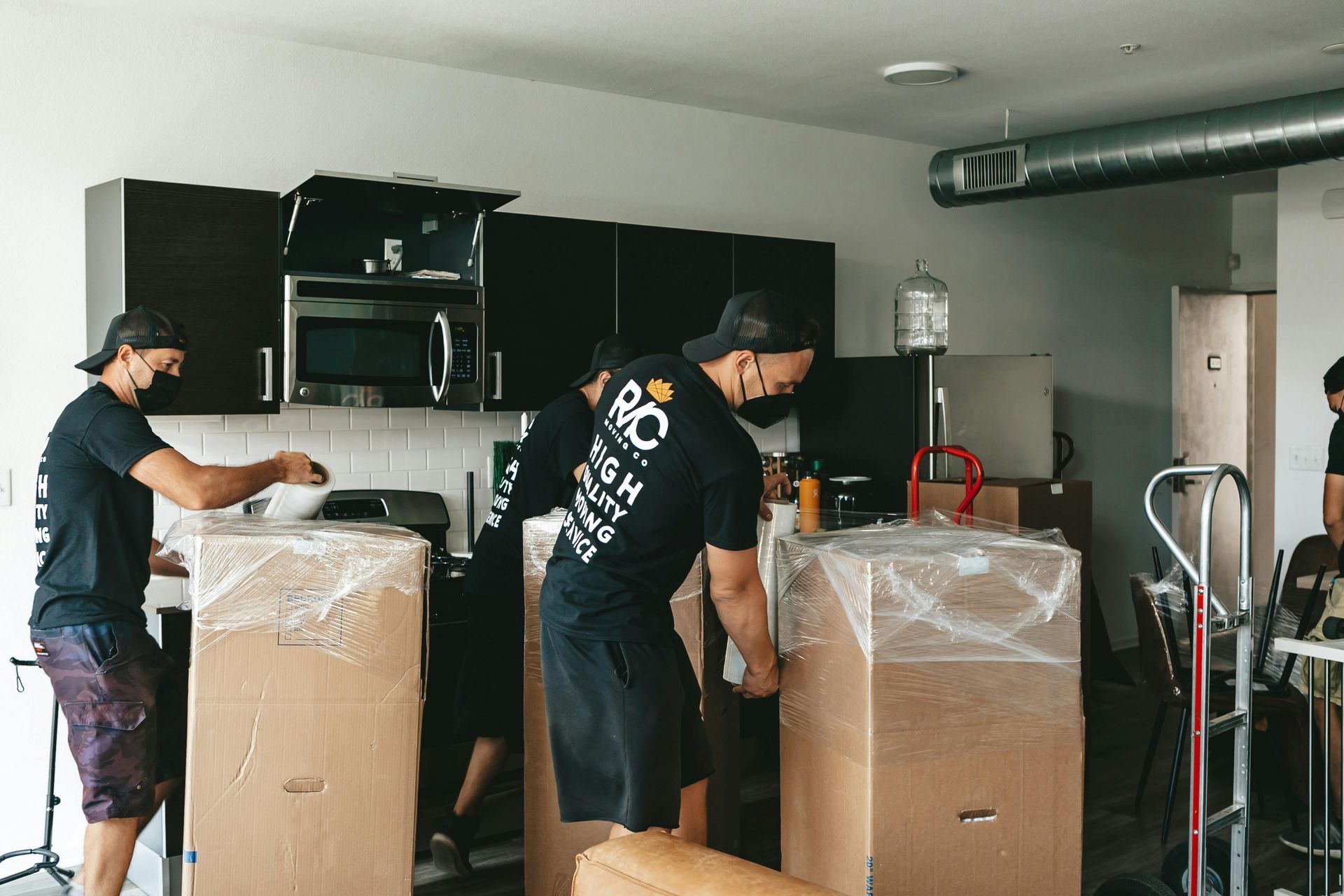 Movers loading boxes in a kitchen with dark cabinets; wearing masks and black shirts.