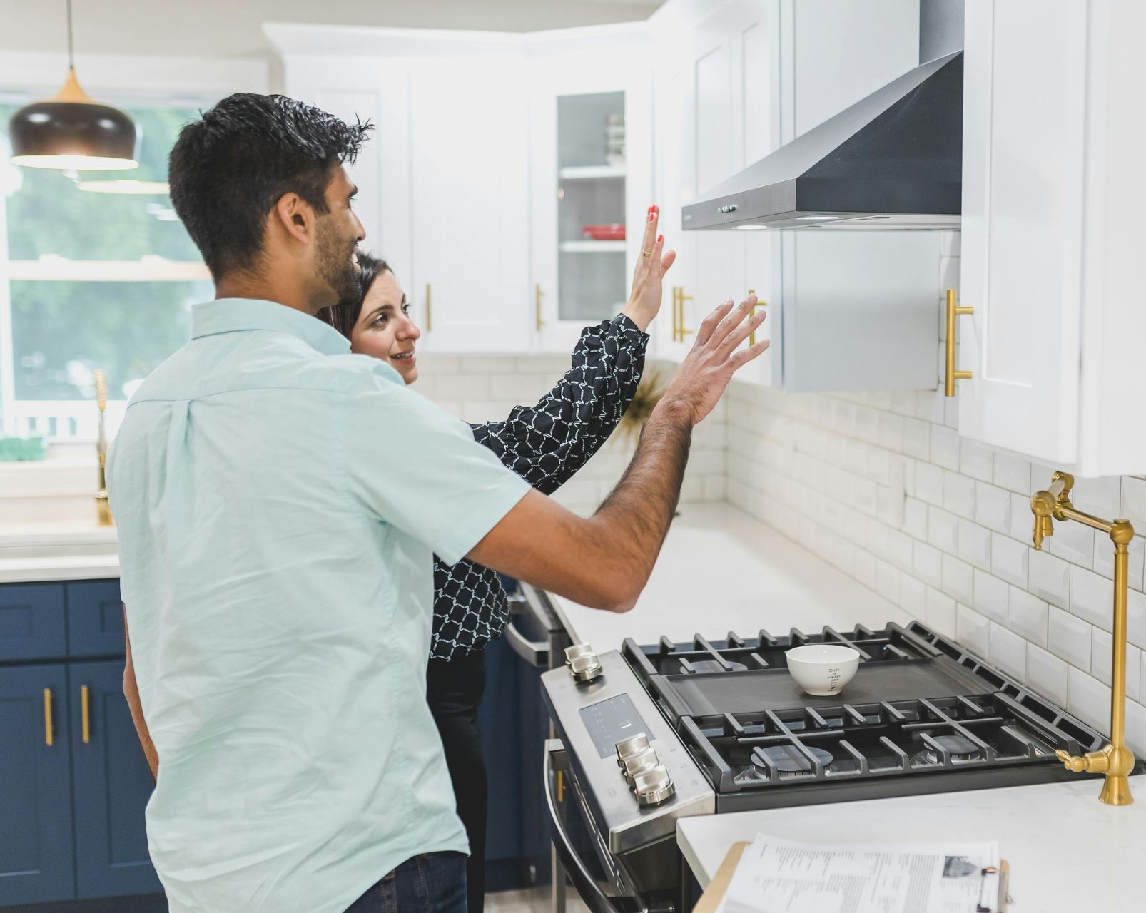 Couple in a white kitchen examining cabinets above a gas stove; one person points.