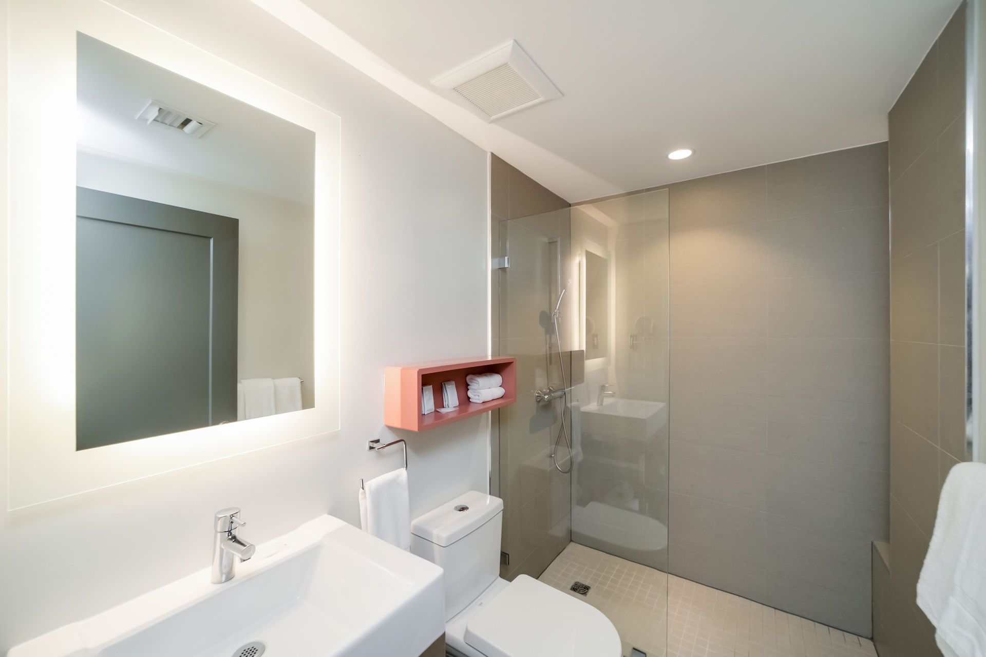 Modern bathroom with a white sink, toilet, and shower. A mirror is above the sink, and a pink shelf is on the wall.