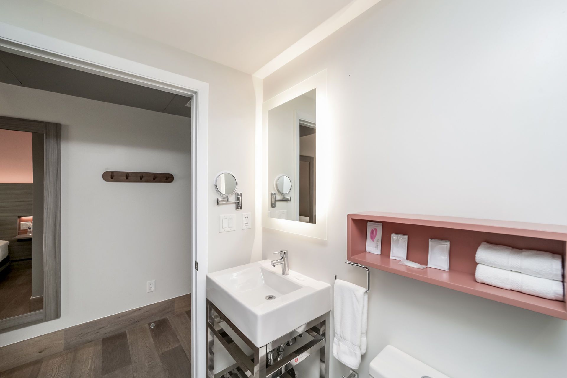 Modern bathroom with a white sink, mirror, and pink shelving. Doorway to a bedroom is visible.