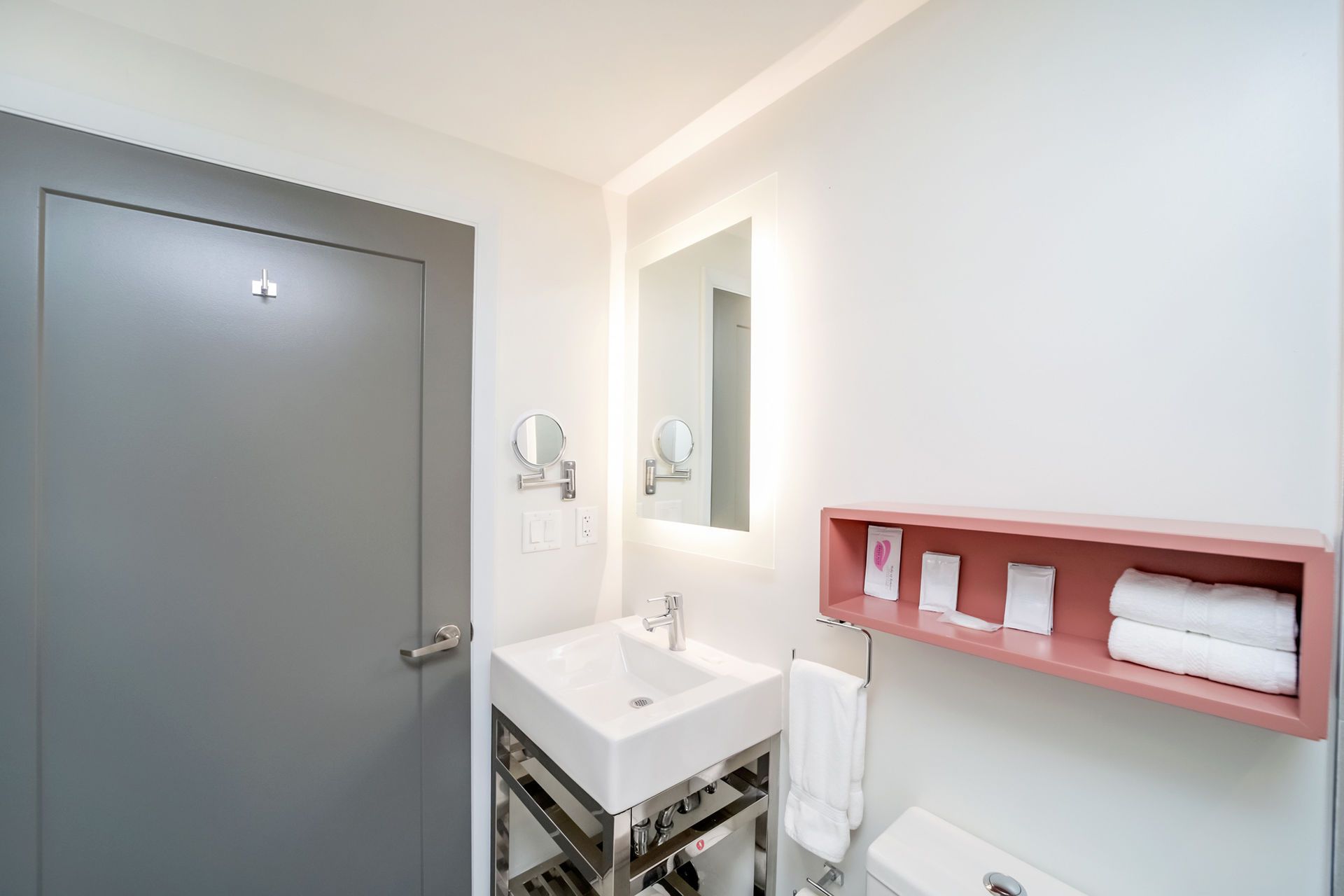 Bathroom with white walls, a gray door, square sink, illuminated mirror, and pink shelf with towels.