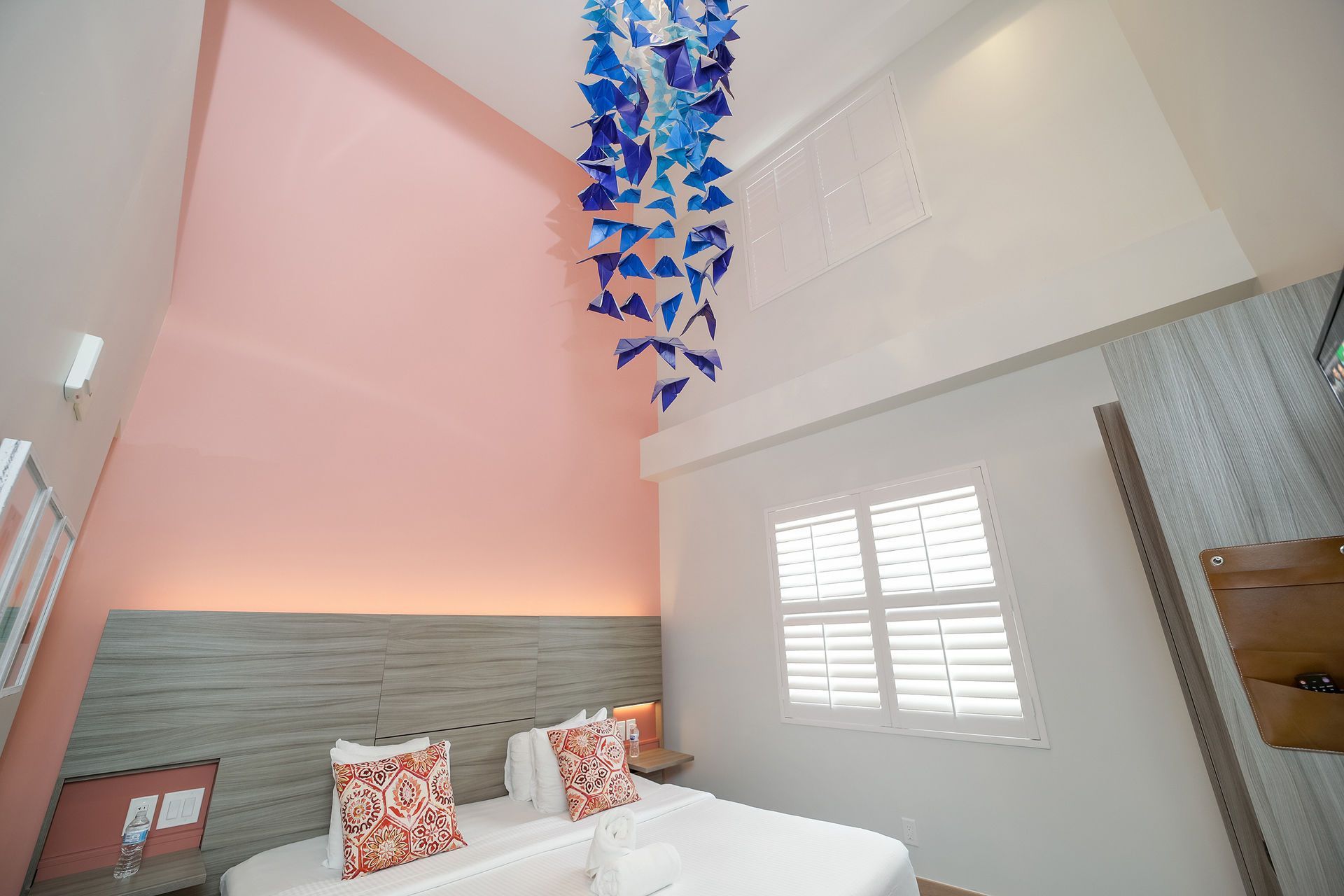 A bedroom with a high pink wall, a blue hanging mobile, a light wood headboard, and a bed with patterned pillows.