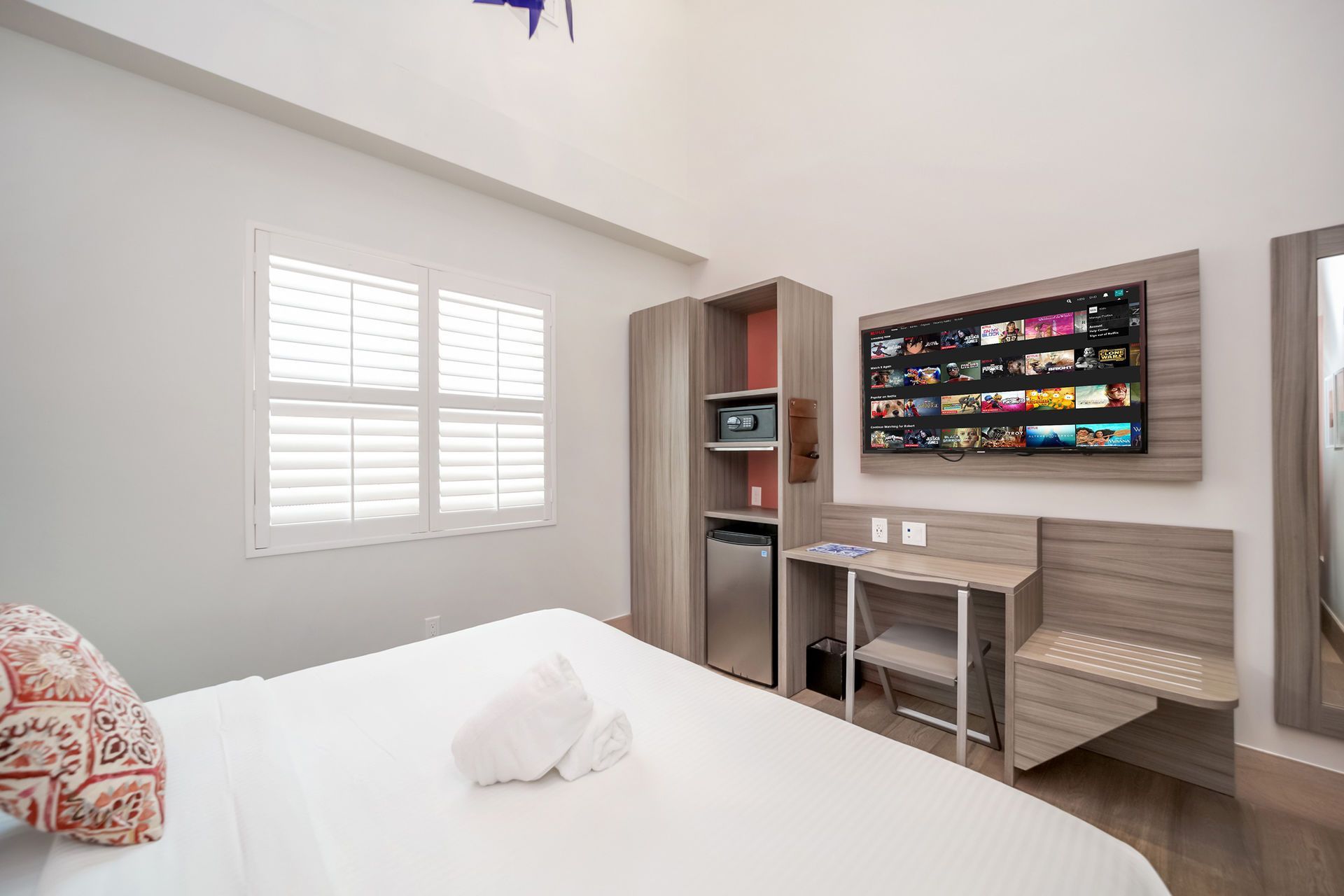 Bedroom with white bedding, TV, window with blinds, desk, and a small fridge.
