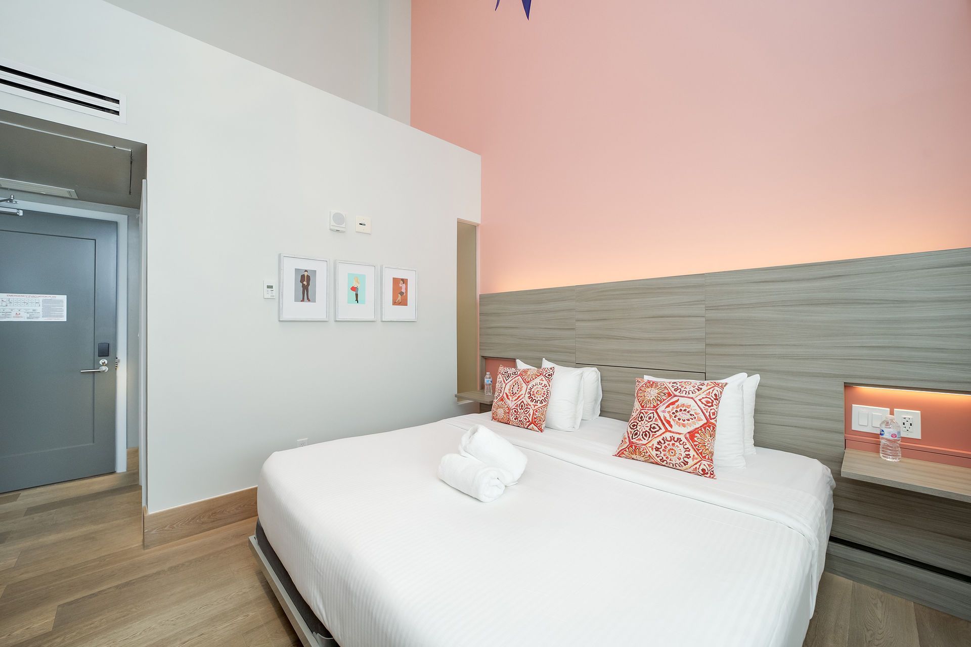 Hotel bedroom with a king-sized bed, decorative pillows, light wood floors, and a pink accent wall.