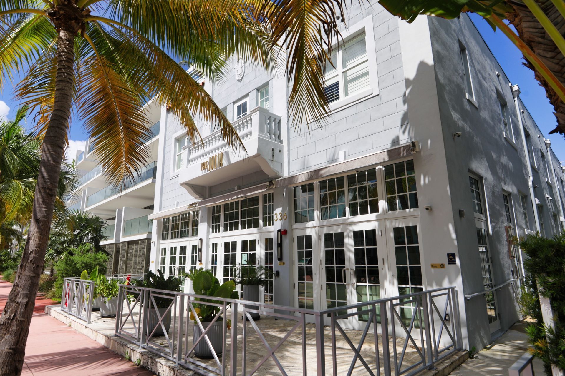 Exterior view of a light gray building with large glass doors and windows, palm trees, and a sidewalk.
