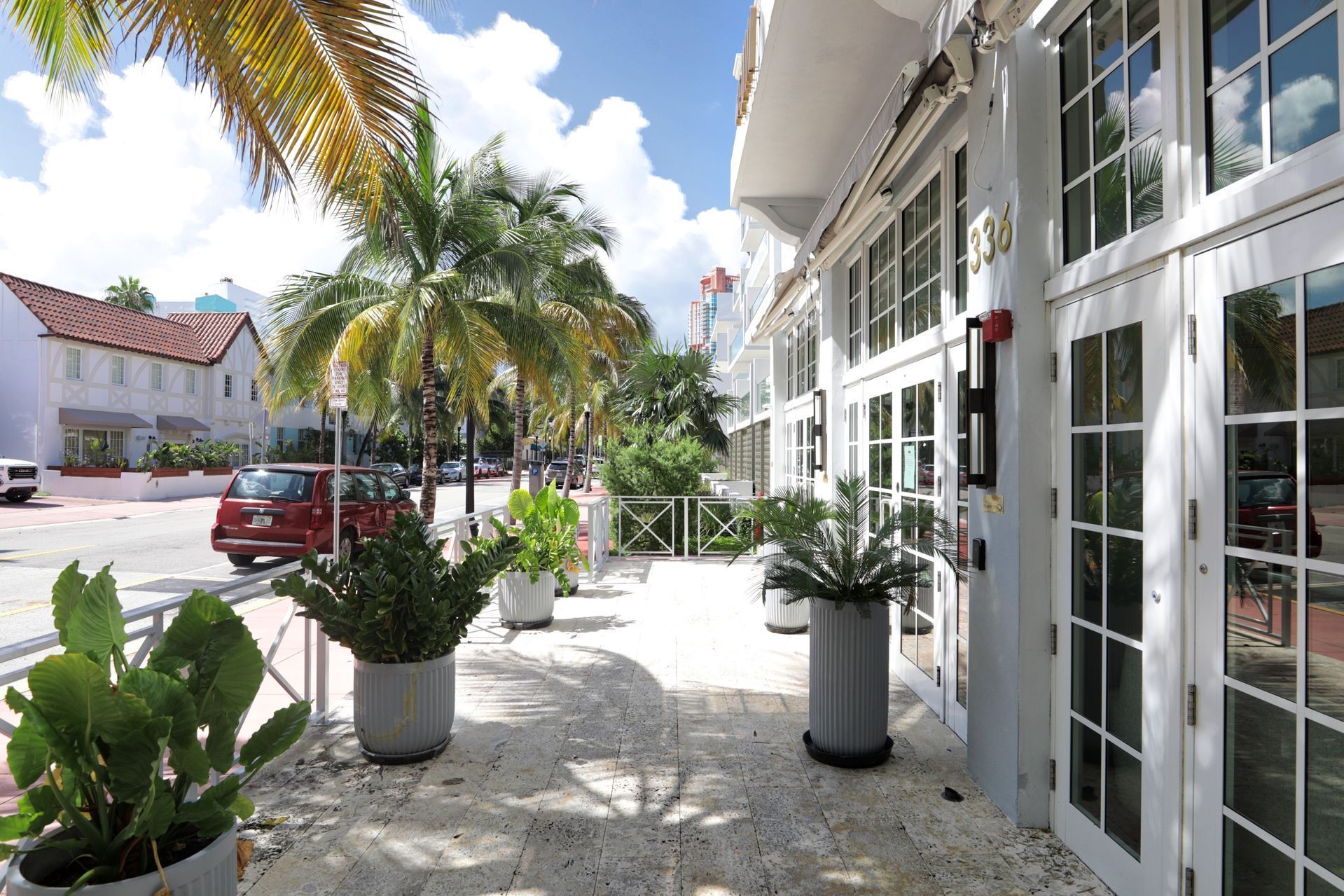 Sidewalk with potted plants, palm trees, and building with white doors. Cars on street. Sunny day.