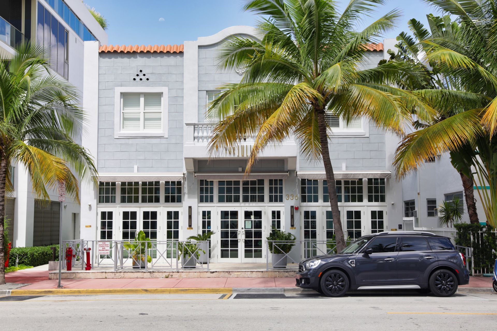 Exterior view of a light gray building with white doors, windows, and a small balcony. A black car is parked in front.