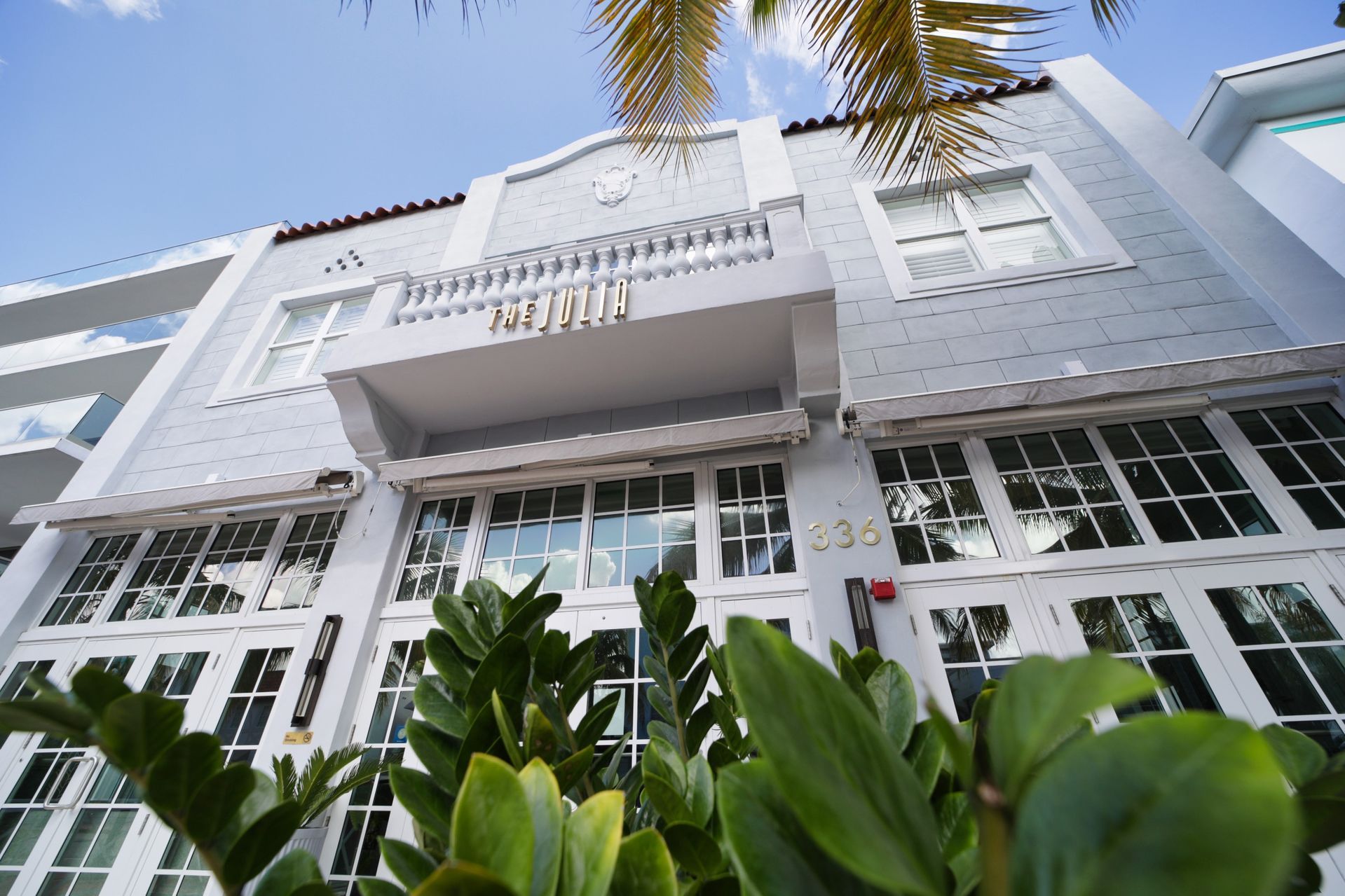 Gray building with white trim, arched facade, balcony, and large windows. Palm fronds in the upper right.