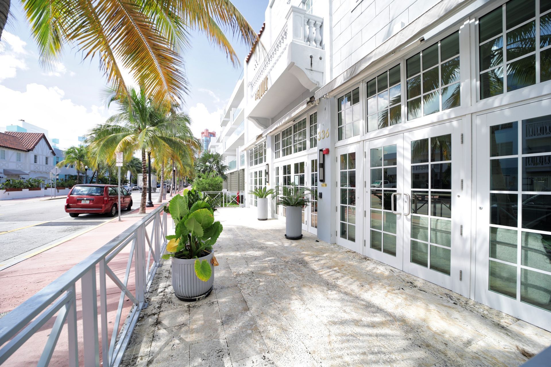 Sidewalk with white building and glass doors, palm trees, red car.