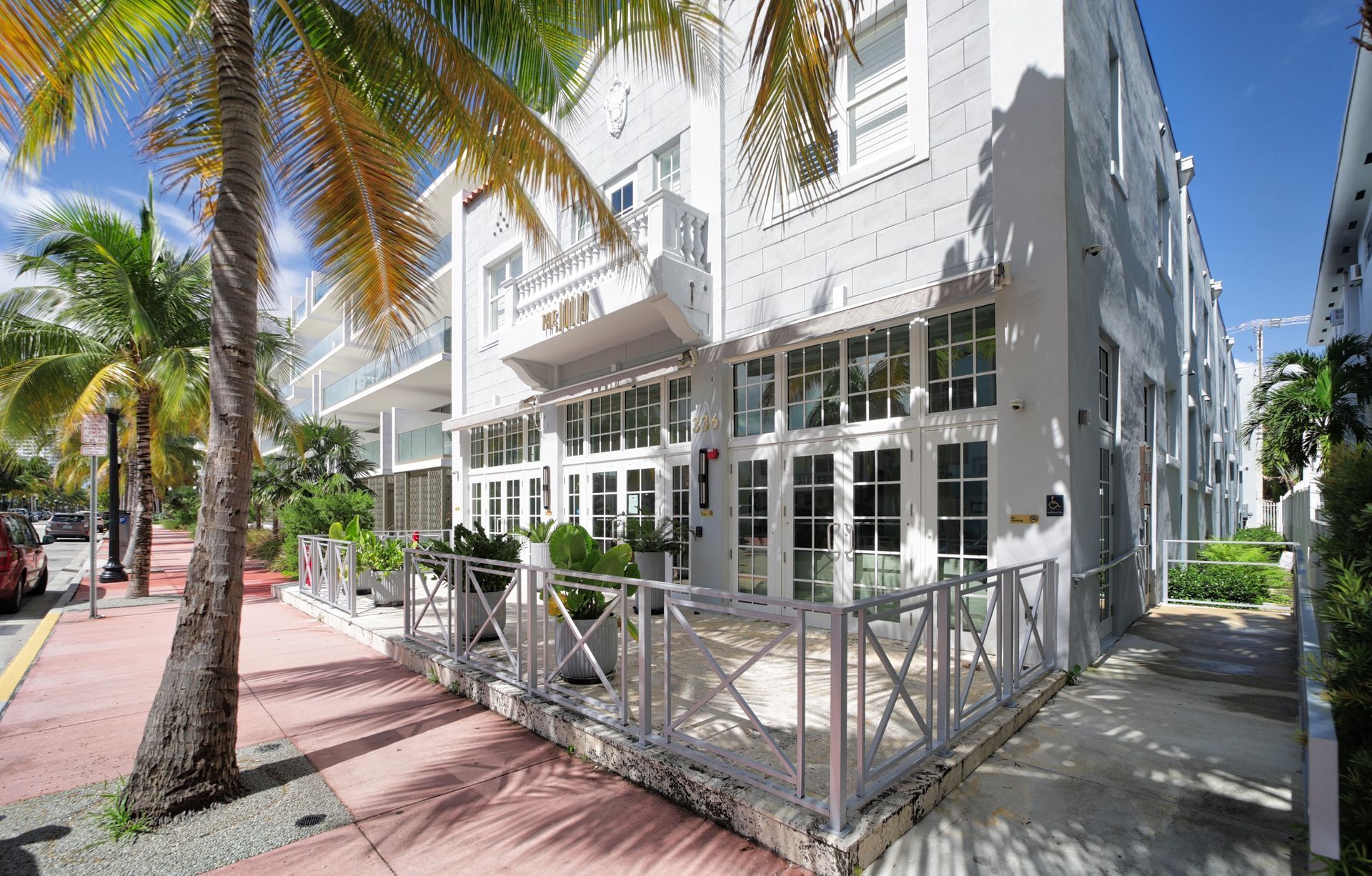 Exterior of a white art deco building with a sidewalk, palm trees, and outdoor seating.