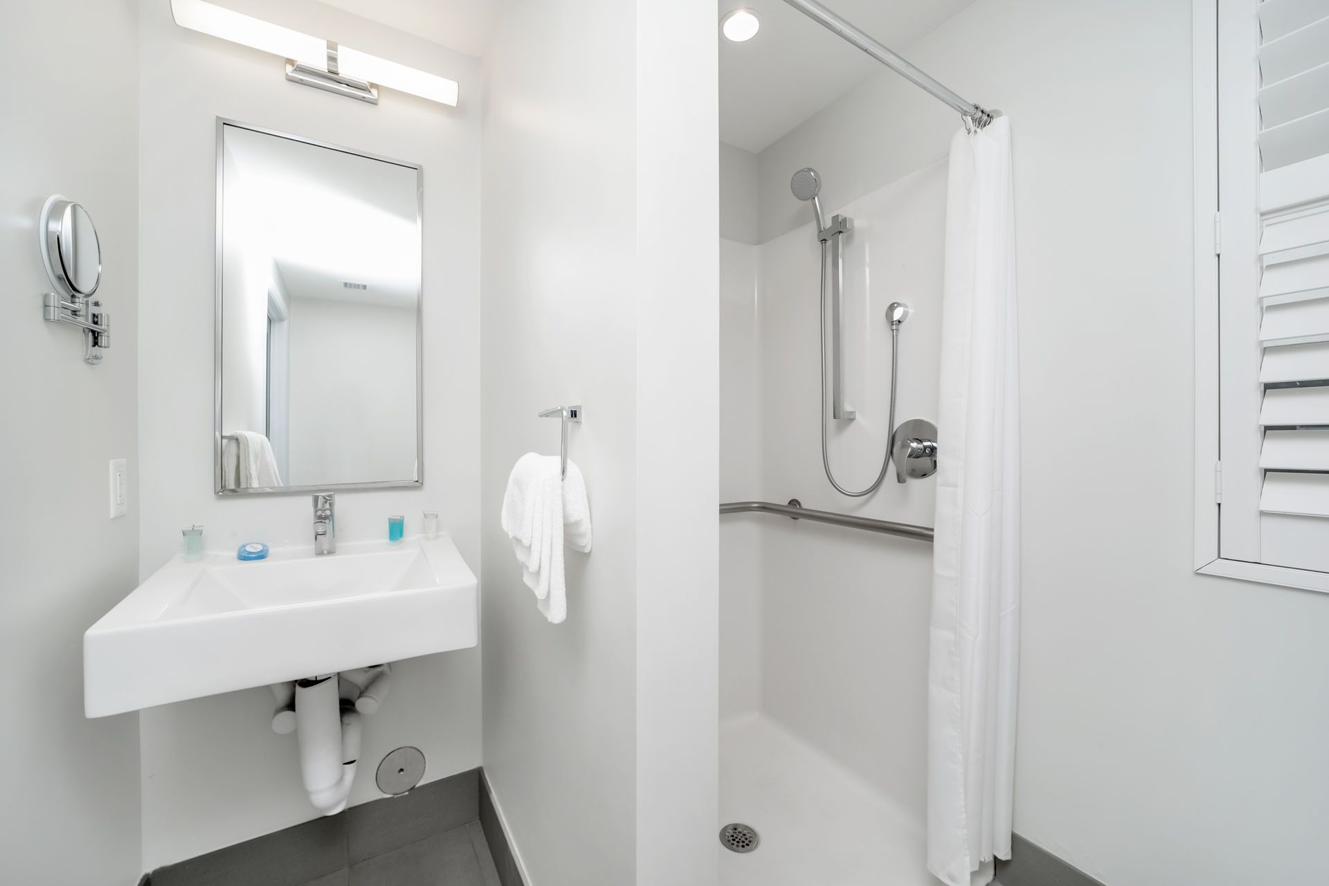 Bright white accessible bathroom with a sink, shower, and window with shutters.