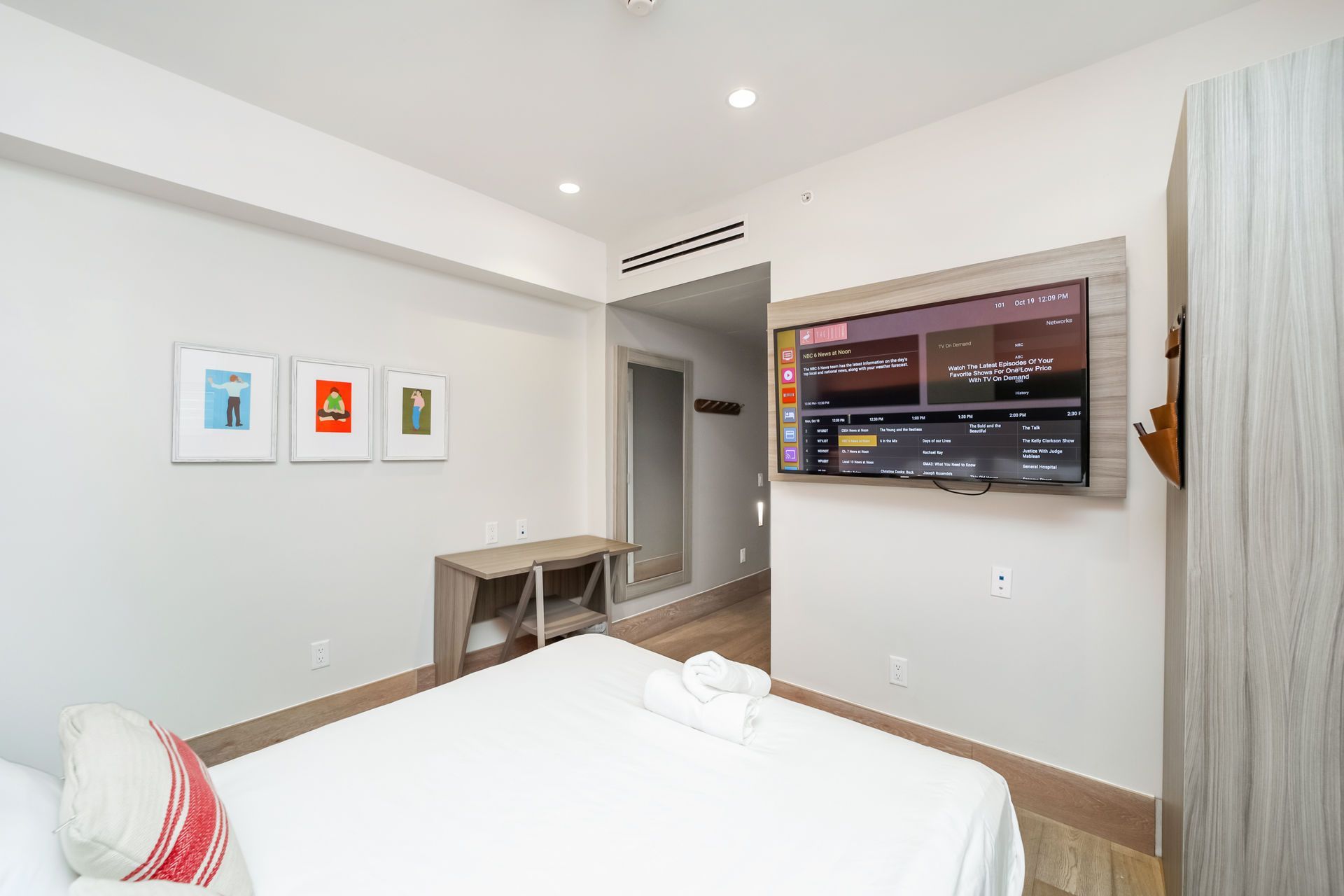 Bedroom with bed, TV, artwork, and a doorway leading to a mirror and hallway. White walls and wood trim.