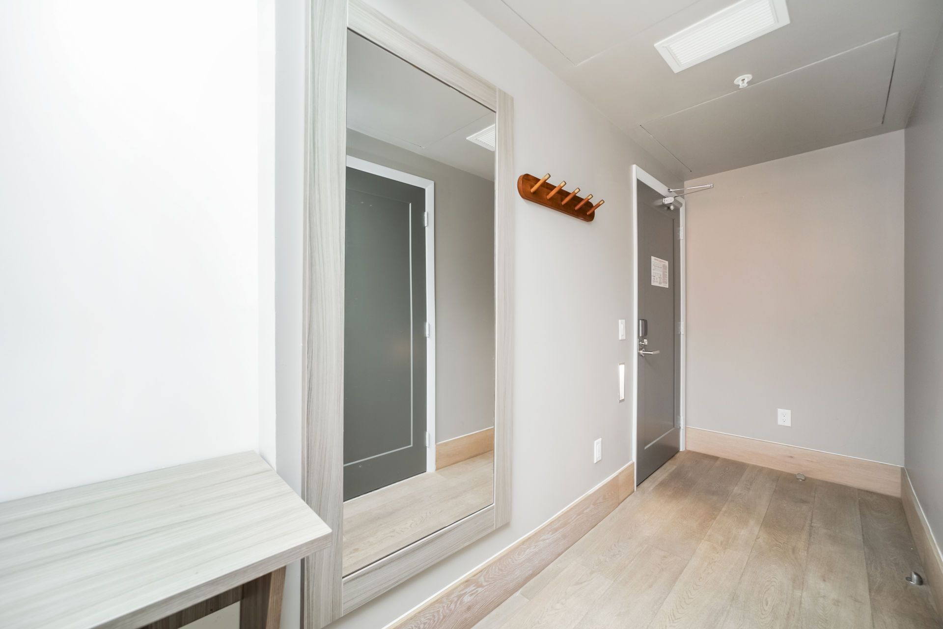 Narrow hallway with full-length mirror, light wood floors, and gray walls. A small bench and coat rack are present.