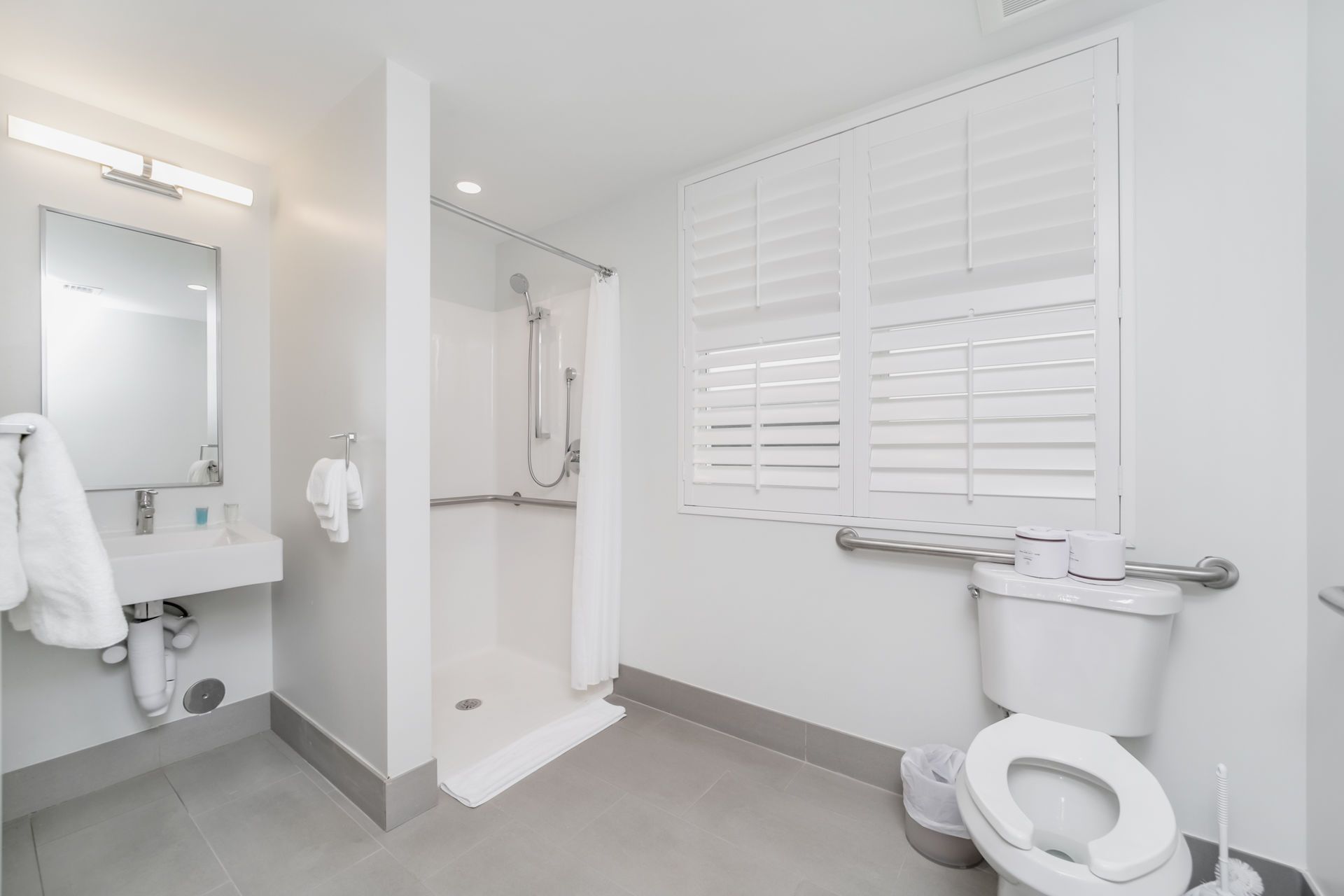 Accessible bathroom with walk-in shower, toilet with grab bar, sink, and shuttered window. White and gray tones.