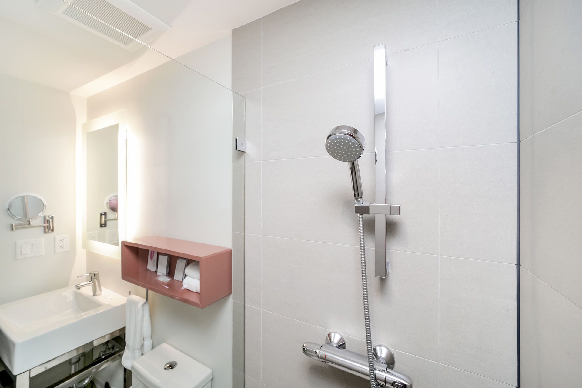 Bathroom with a shower, sink, and toilet. White walls and fixtures; pink shelf.