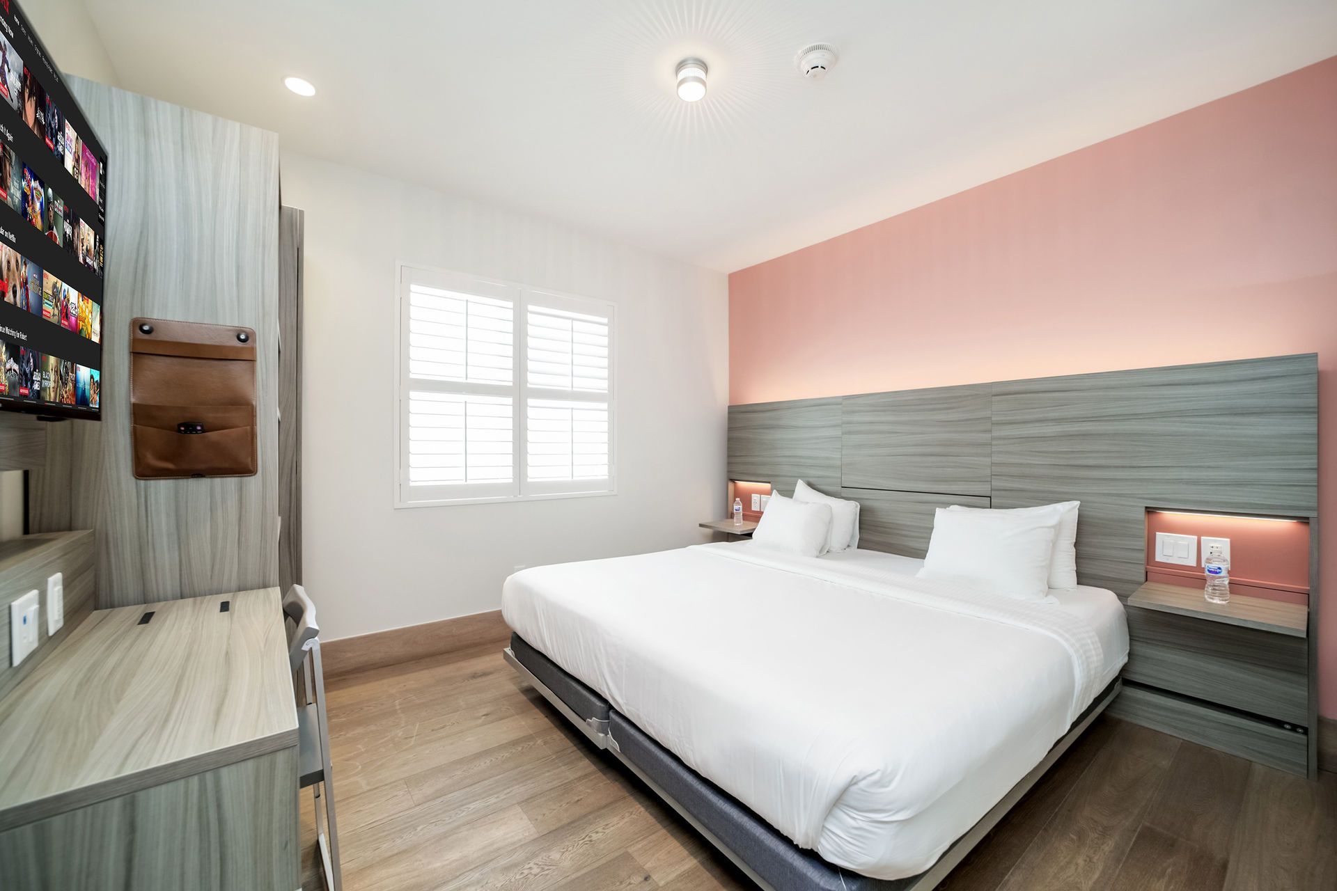 Hotel room with a bed, TV, and window. Pink wall, gray headboard and furniture.