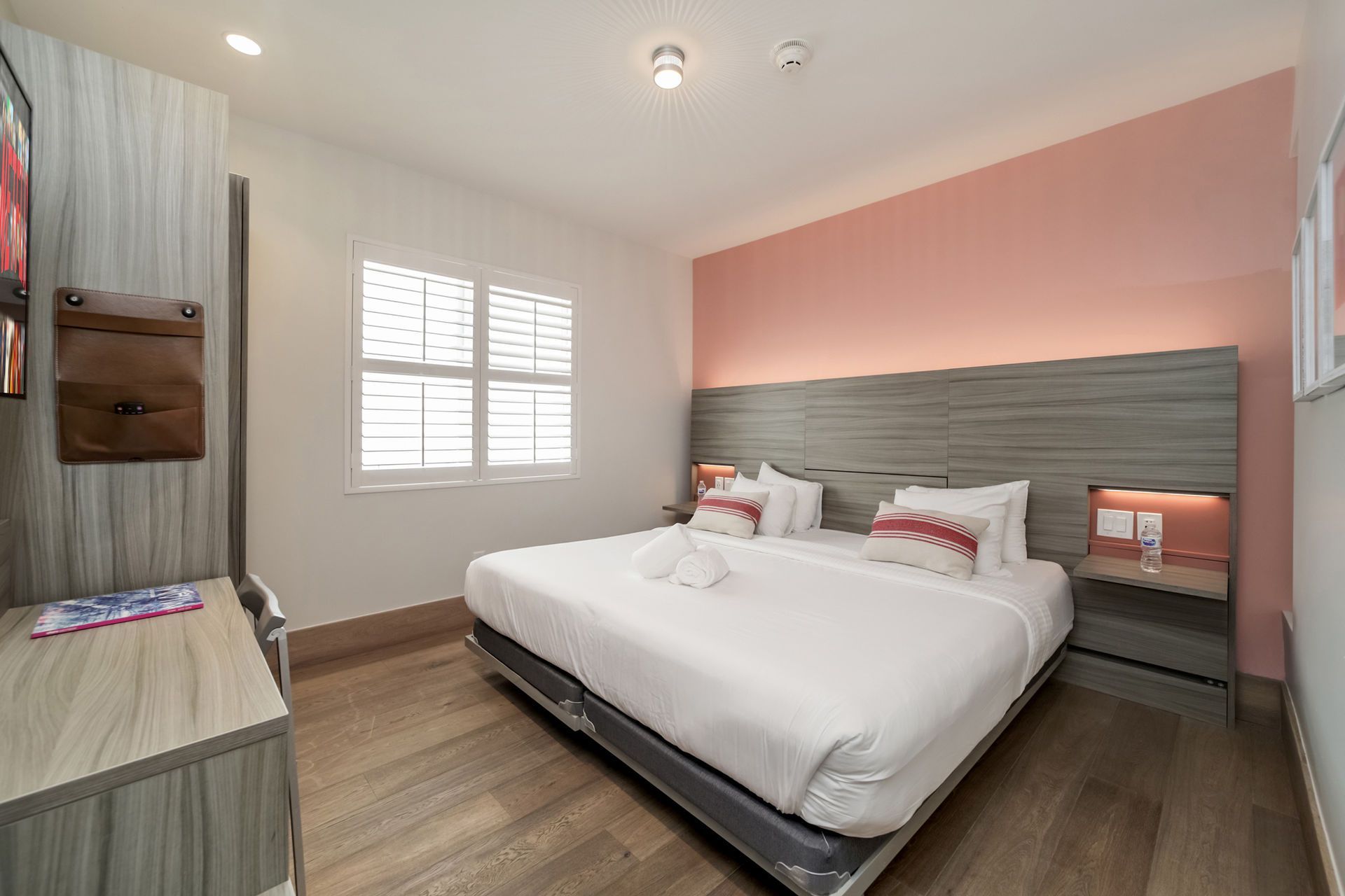 Hotel bedroom with a bed, nightstands, a desk, and a window. The wall behind the bed is pink.