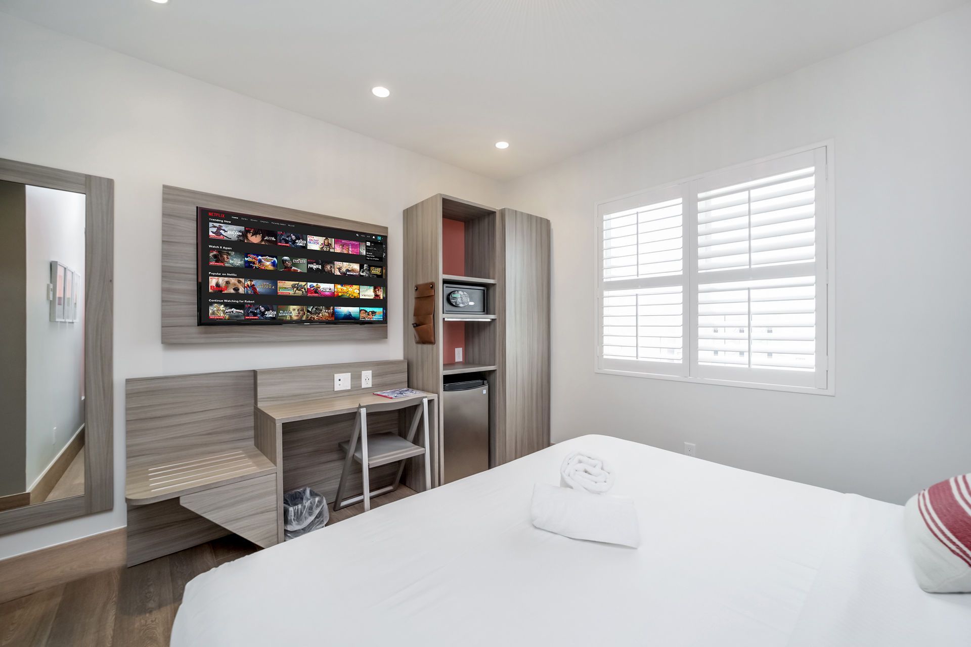 A small hotel room with a bed, TV, desk, and window with shutters.