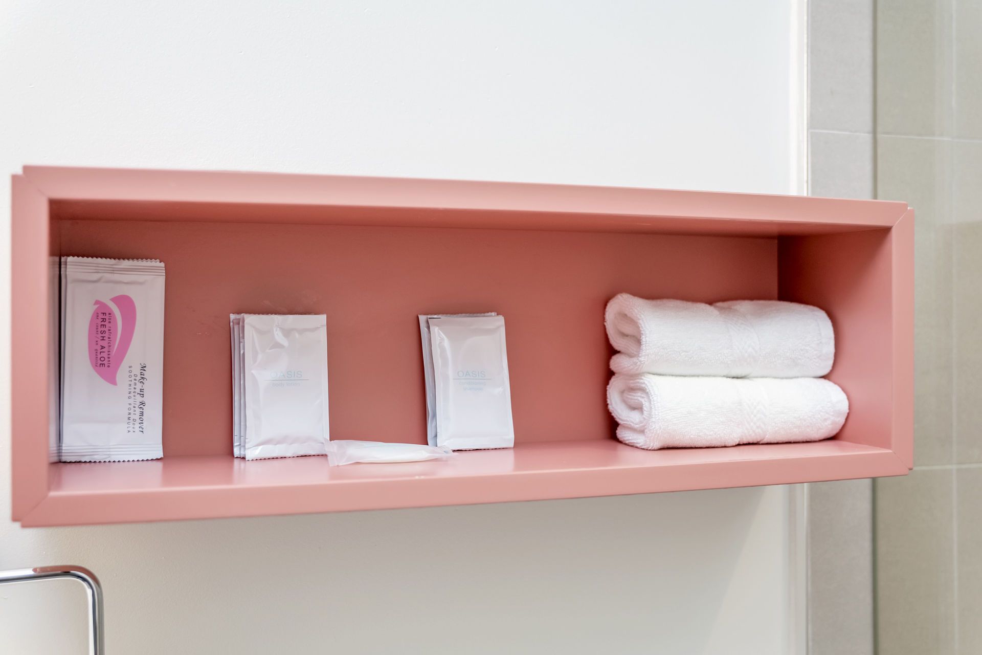Pink shelf built into a wall, holding white-wrapped toiletries and folded white towels.