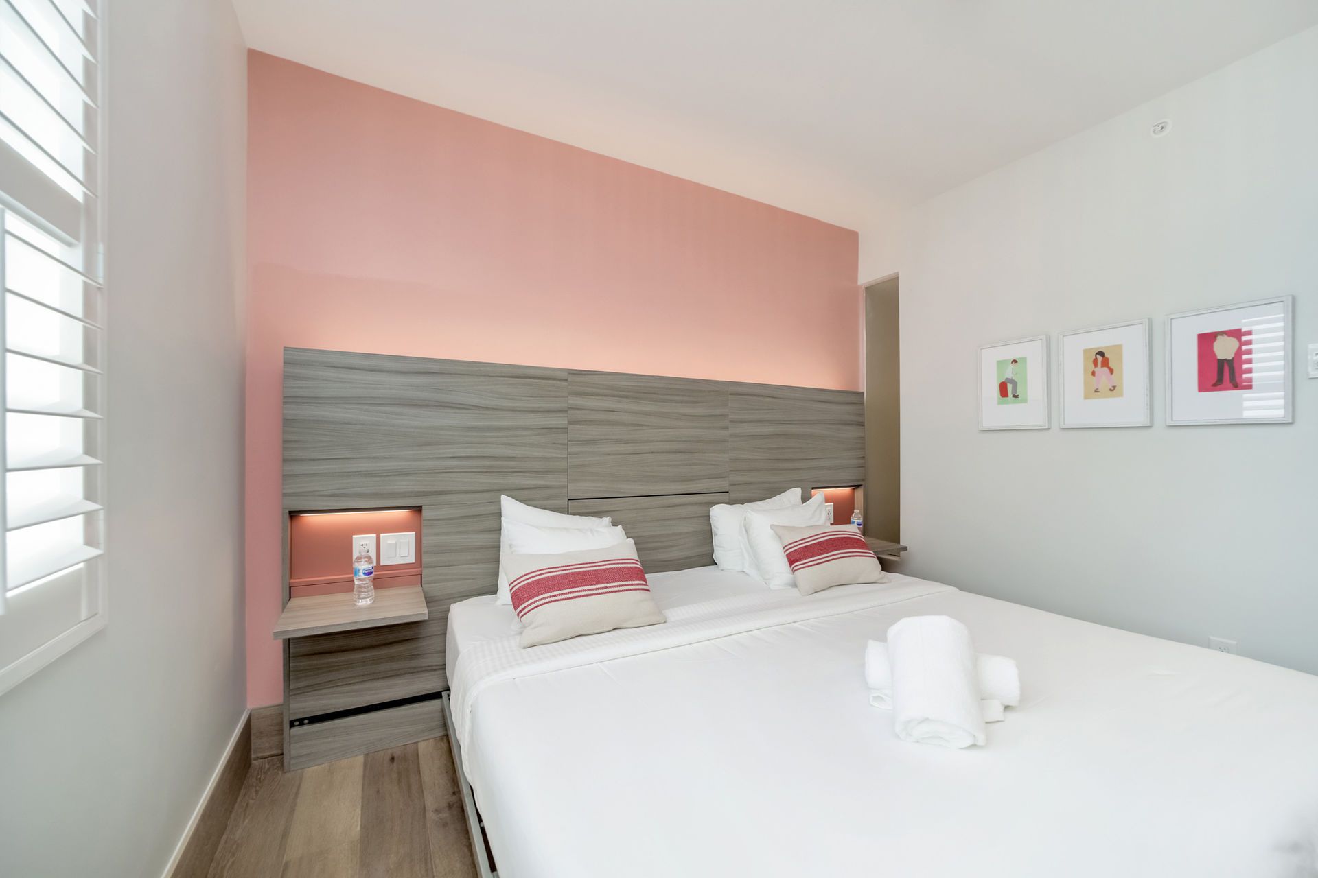 Bedroom with a white bed, pink wall, and artwork. A window with shutters is on the left.