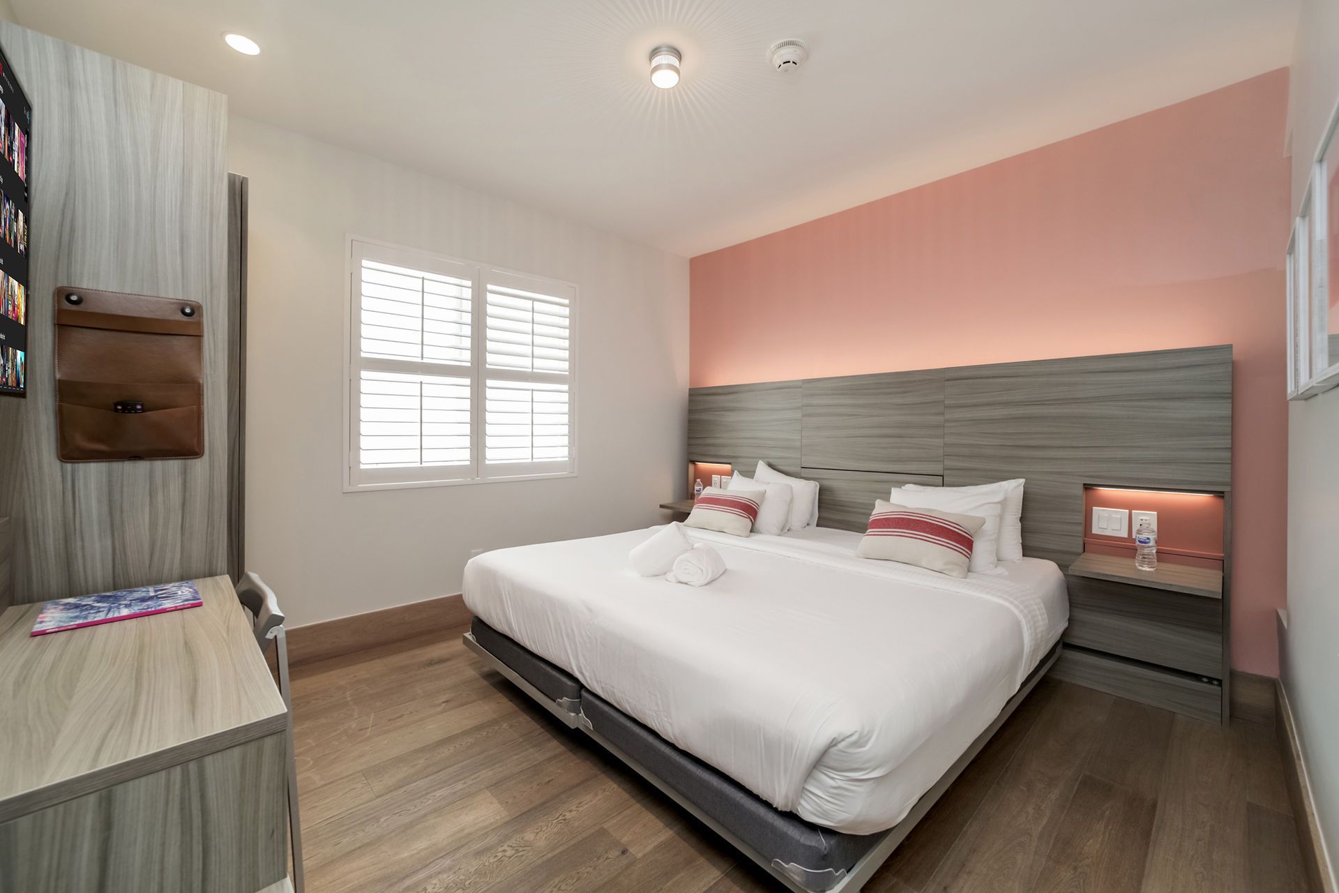 A modern bedroom with light wood flooring, a king bed with white linens, a pink accent wall, and a wooden headboard.
