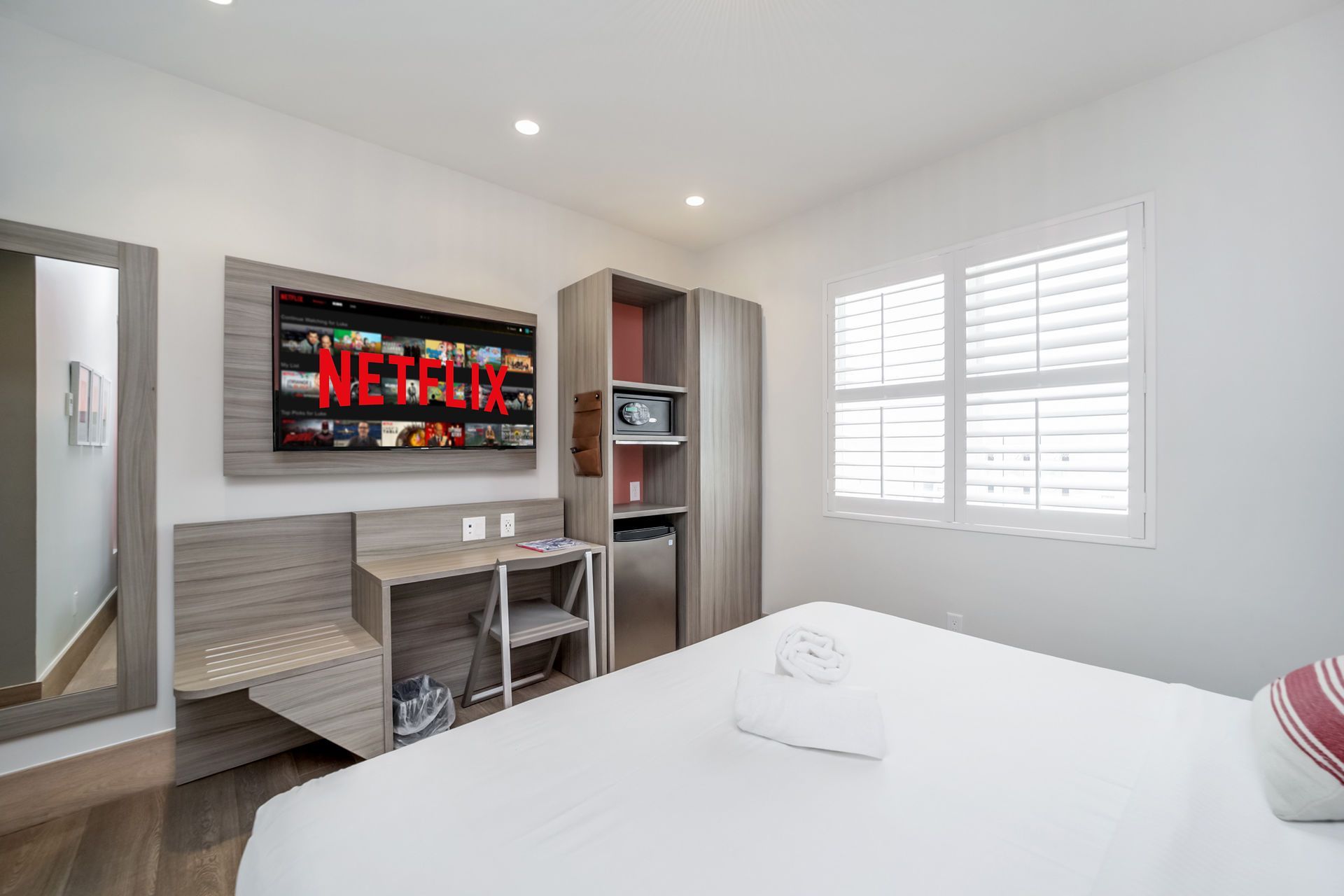 A bright hotel room with a bed, TV, desk, window with blinds, and a storage cabinet.
