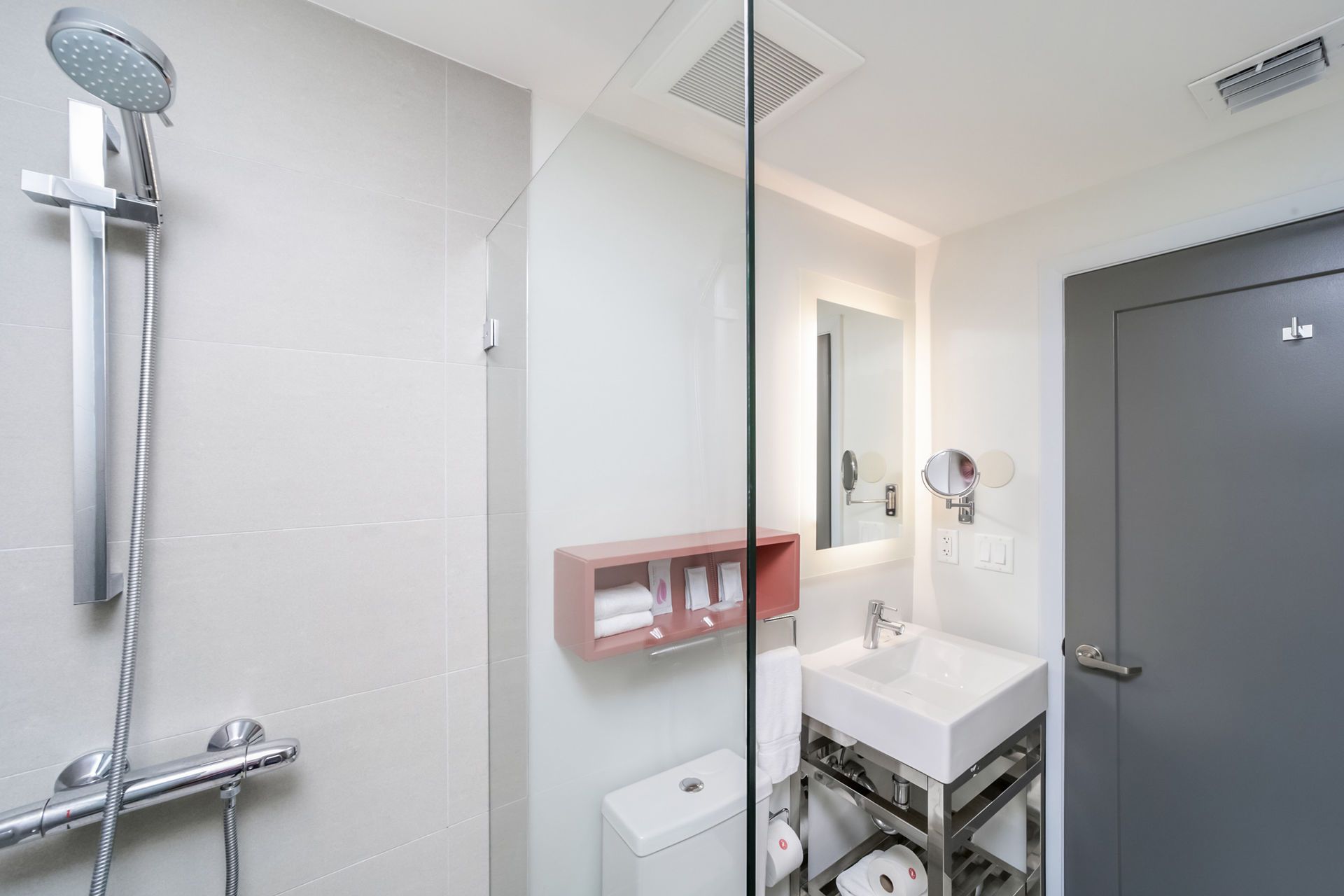 Modern bathroom with shower, toilet, sink, mirror, and a small pink shelf. Gray and white color scheme.