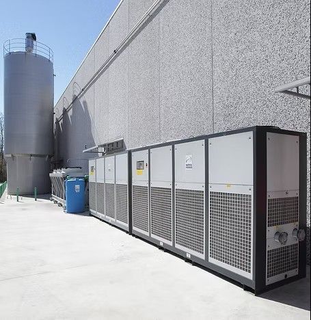 Unità di raffreddamento industriali esterne allineate accanto a un edificio grigio con un silo, in una giornata di sole.