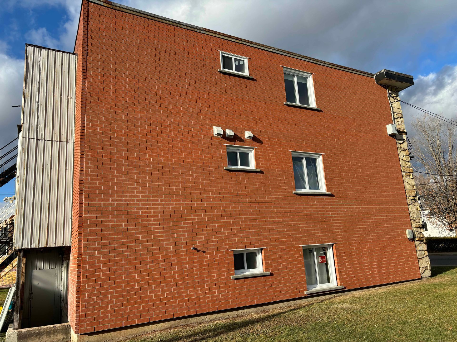 Bâtiment en briques rouges avec de nombreuses fenêtres sur une colline herbeuse ; une structure en bois et des éléments en pierre sont visibles.