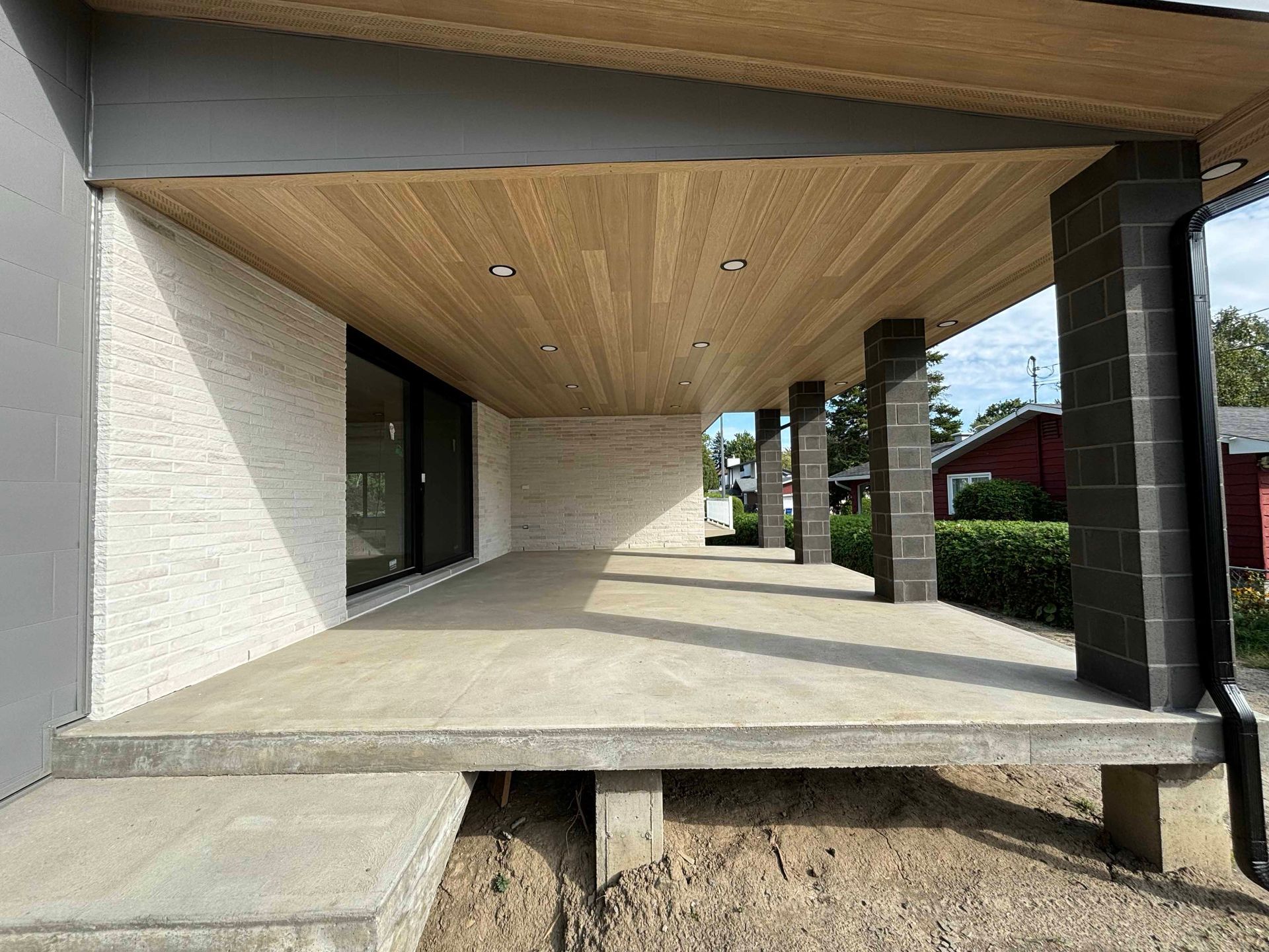 Véranda couverte avec sol en béton, plafond en bois, murs en briques et colonnes de soutien noires.