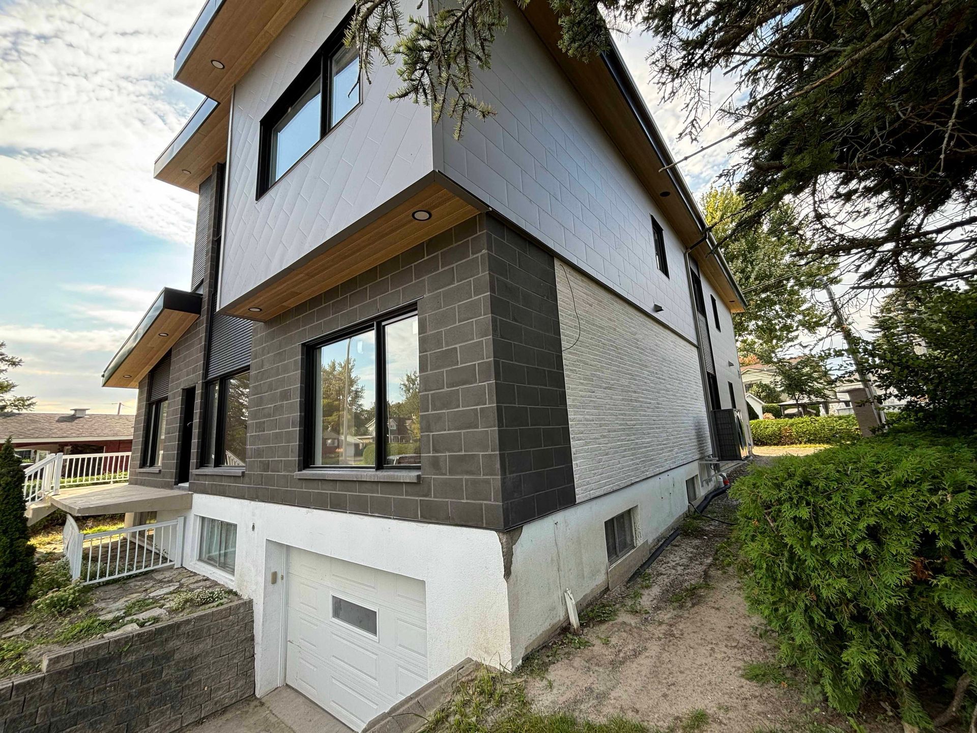 Maison moderne à deux étages avec revêtement extérieur blanc, gris et marron. Garage, fenêtres et aménagement paysager. Journée ensoleillée.
