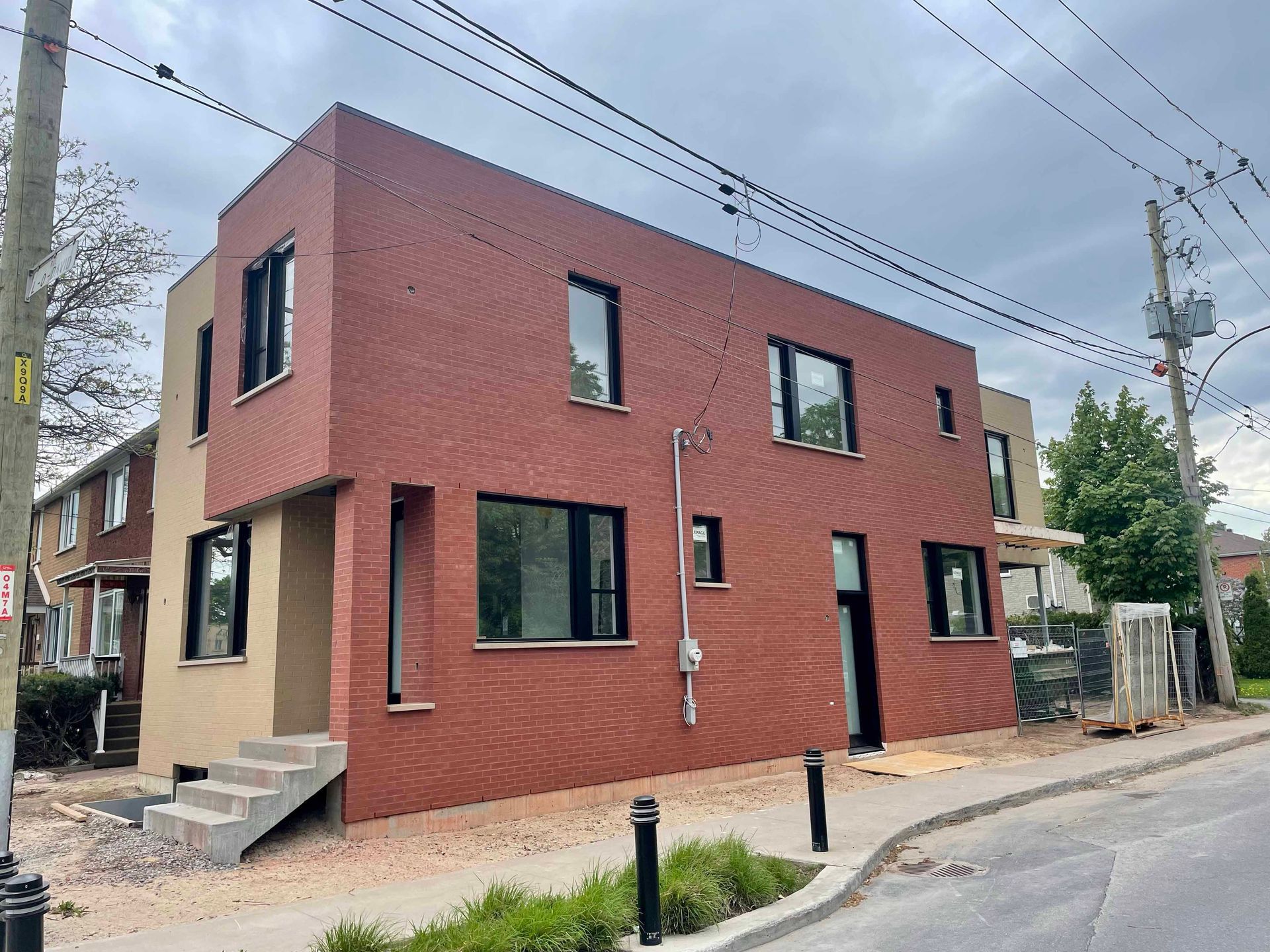 Bâtiment en briques de deux étages avec fenêtres noires, enduit beige et marches en béton ; vue sur la rue.