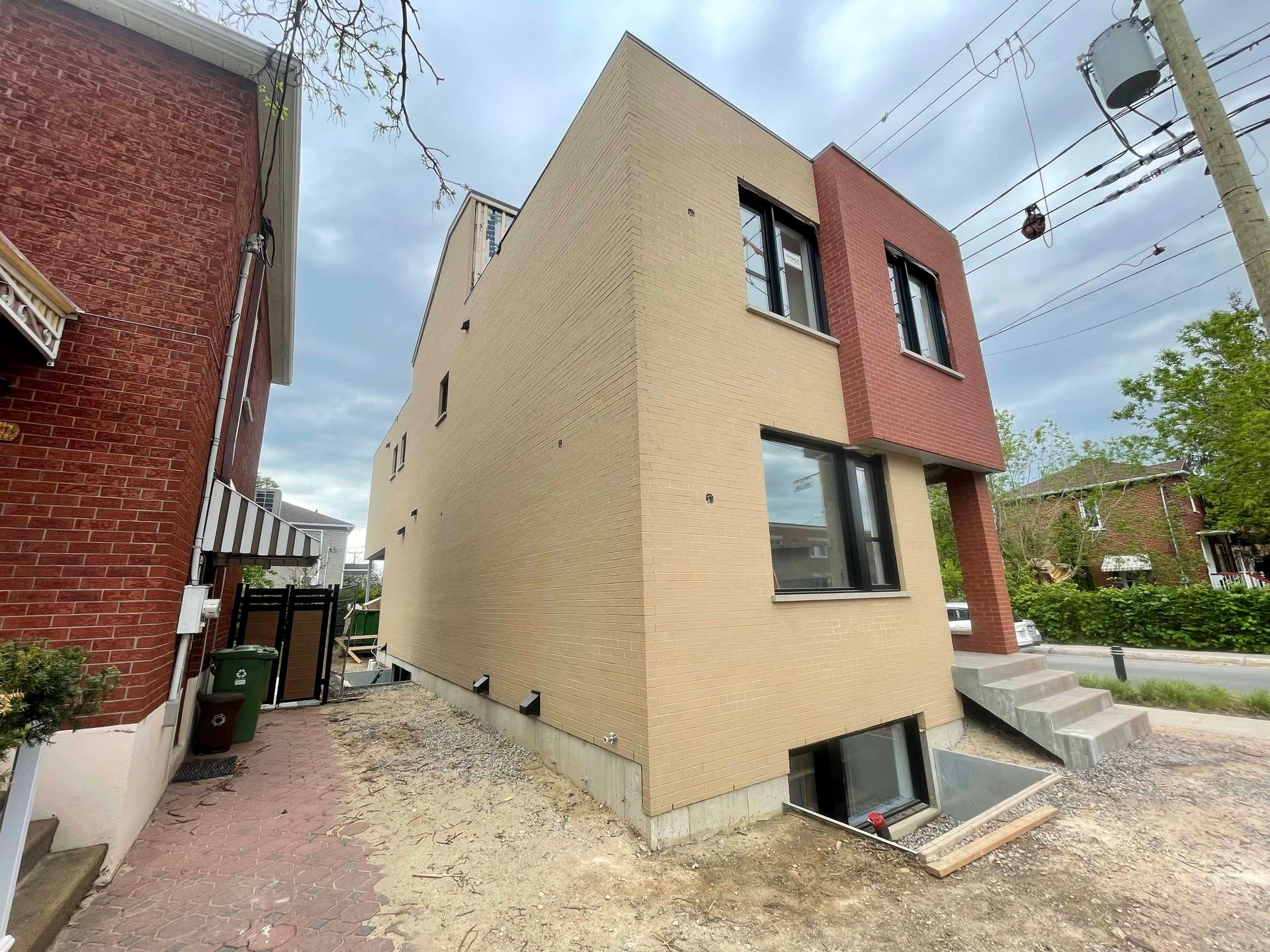 Vue latérale d'un bâtiment de trois étages à façade en briques beiges et rouges, marches menant à la porte, ciel nuageux.