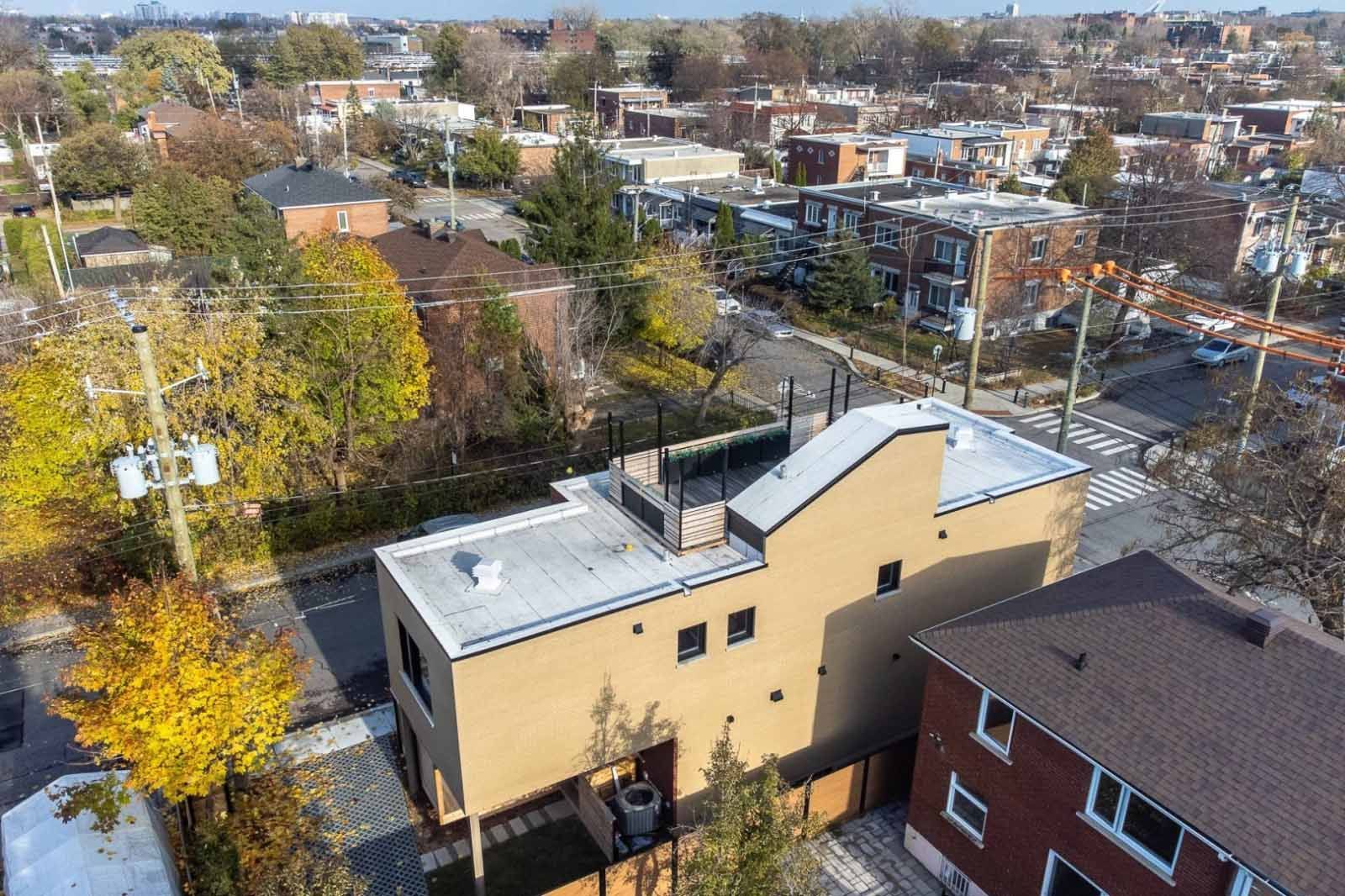 Vue aérienne d'un bâtiment beige de deux étages à toit plat, entouré d'autres bâtiments et d'arbres, par une journée ensoleillée.