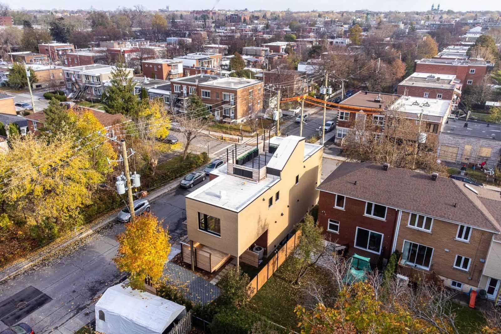 Vue aérienne d'une maison moderne de couleur beige avec un toit plat, entourée d'immeubles résidentiels et d'arbres en milieu urbain.