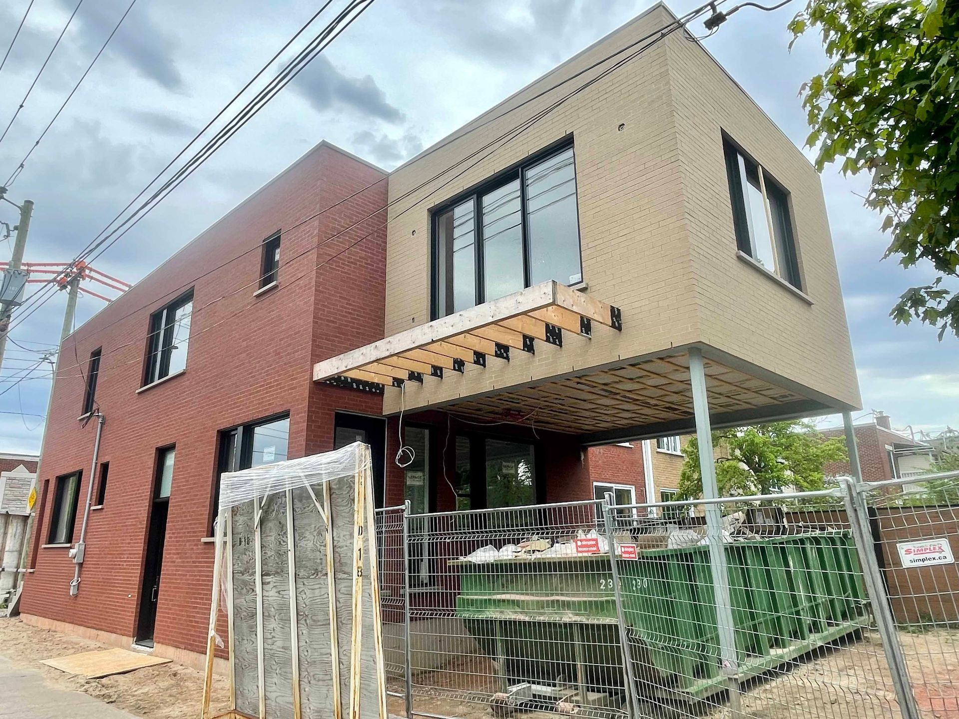 Maison de deux étages en construction, avec des briques rouges et un bardage beige, un balcon et des échafaudages.