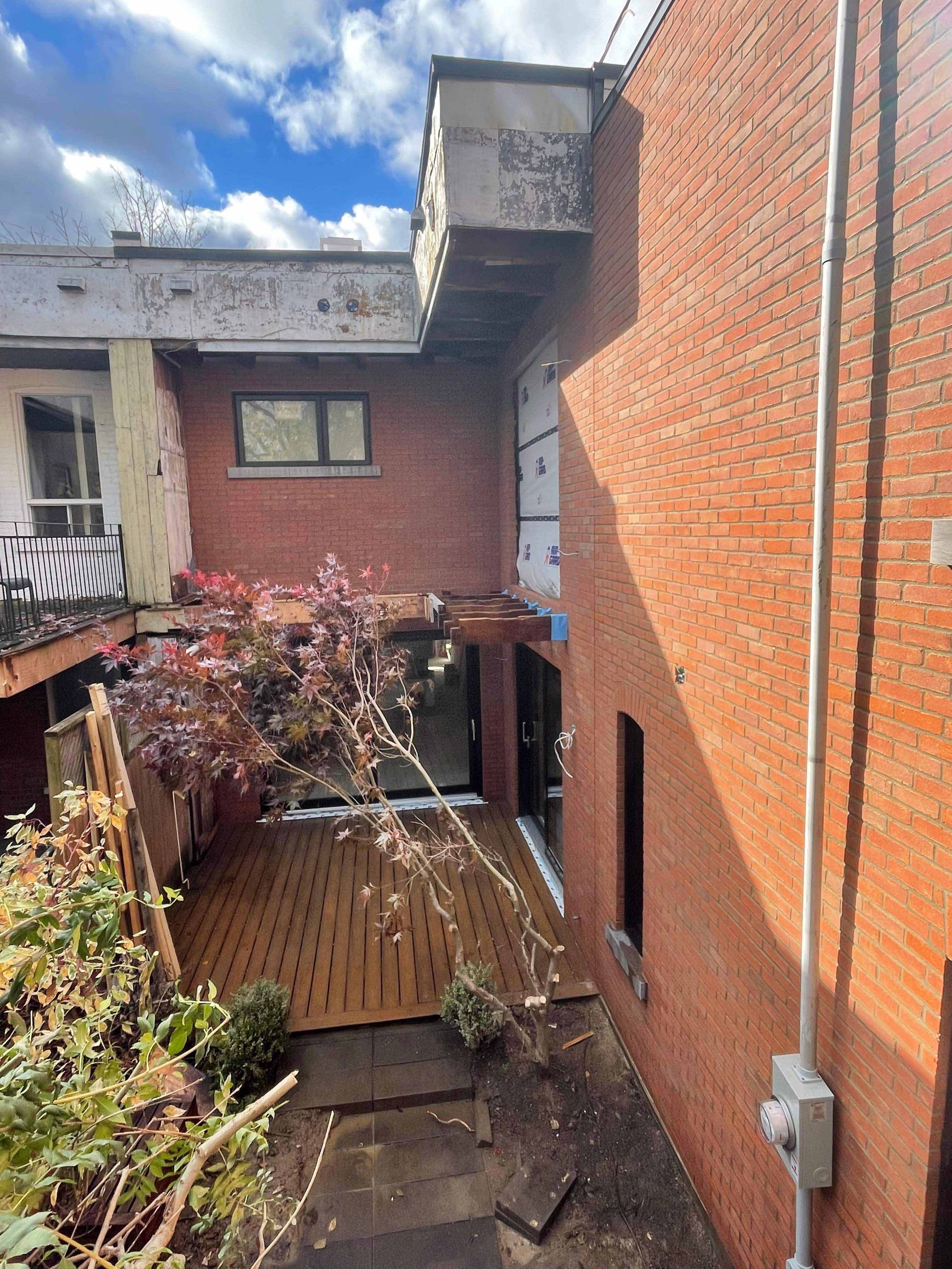 Jardin avec terrasse en bois, dépendances en briques et petit arbre. Journée ensoleillée sous un ciel nuageux.
