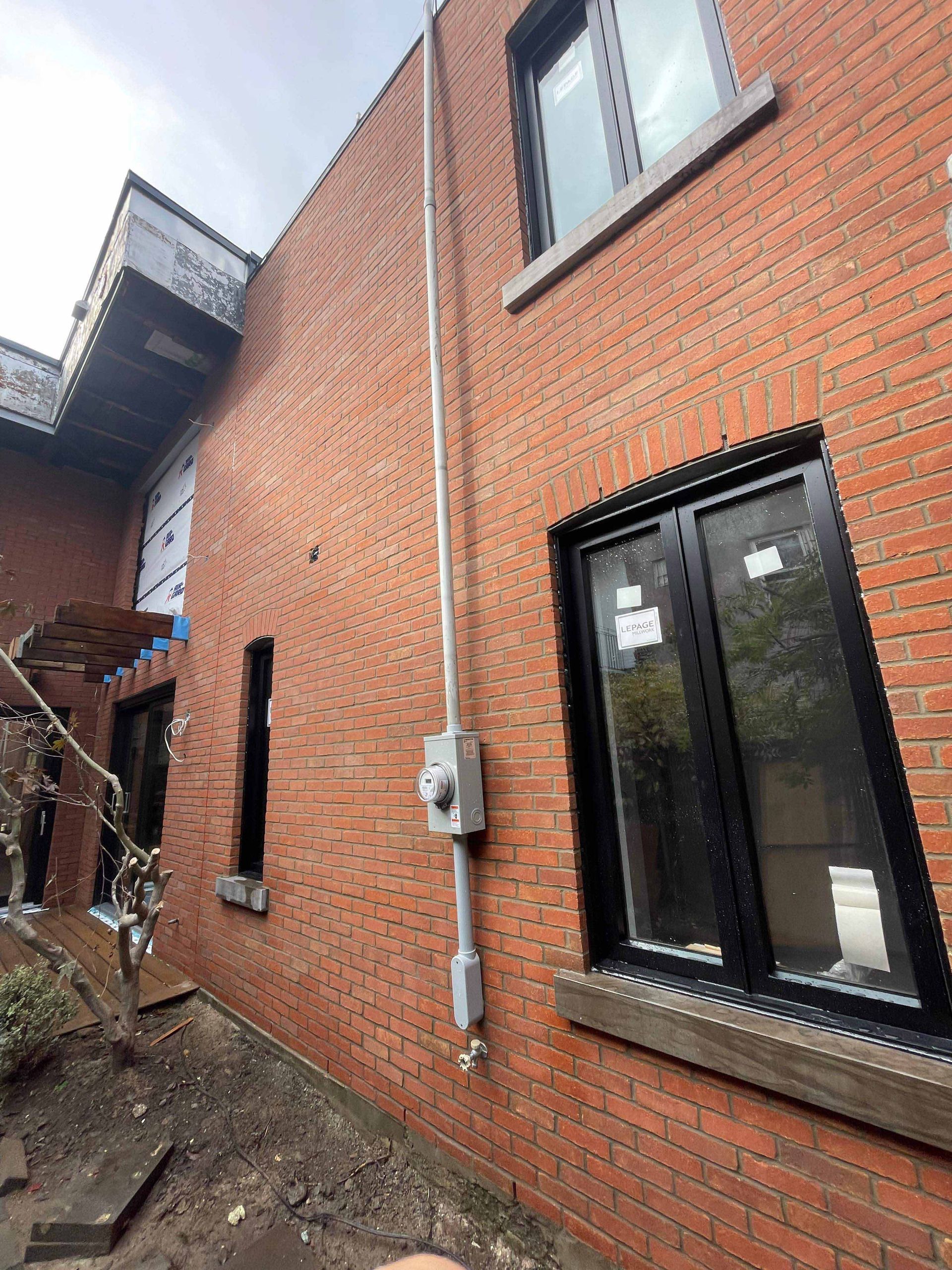 Bâtiment en briques avec conduit électrique et compteur. Fenêtres à cadre noir visibles.