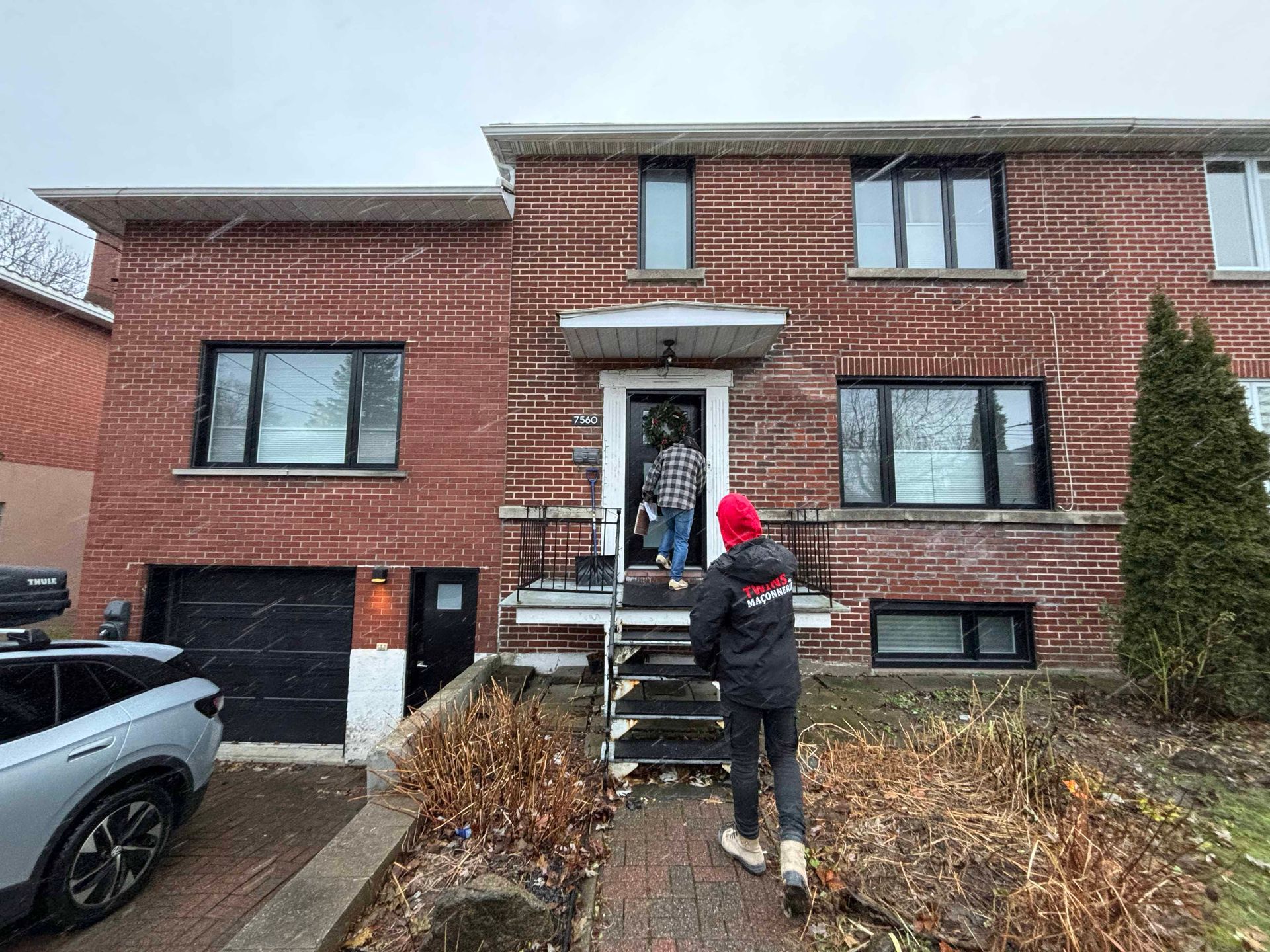 Une personne marche vers une maison en briques, une autre personne se tient devant la porte d'entrée ; journée enneigée.