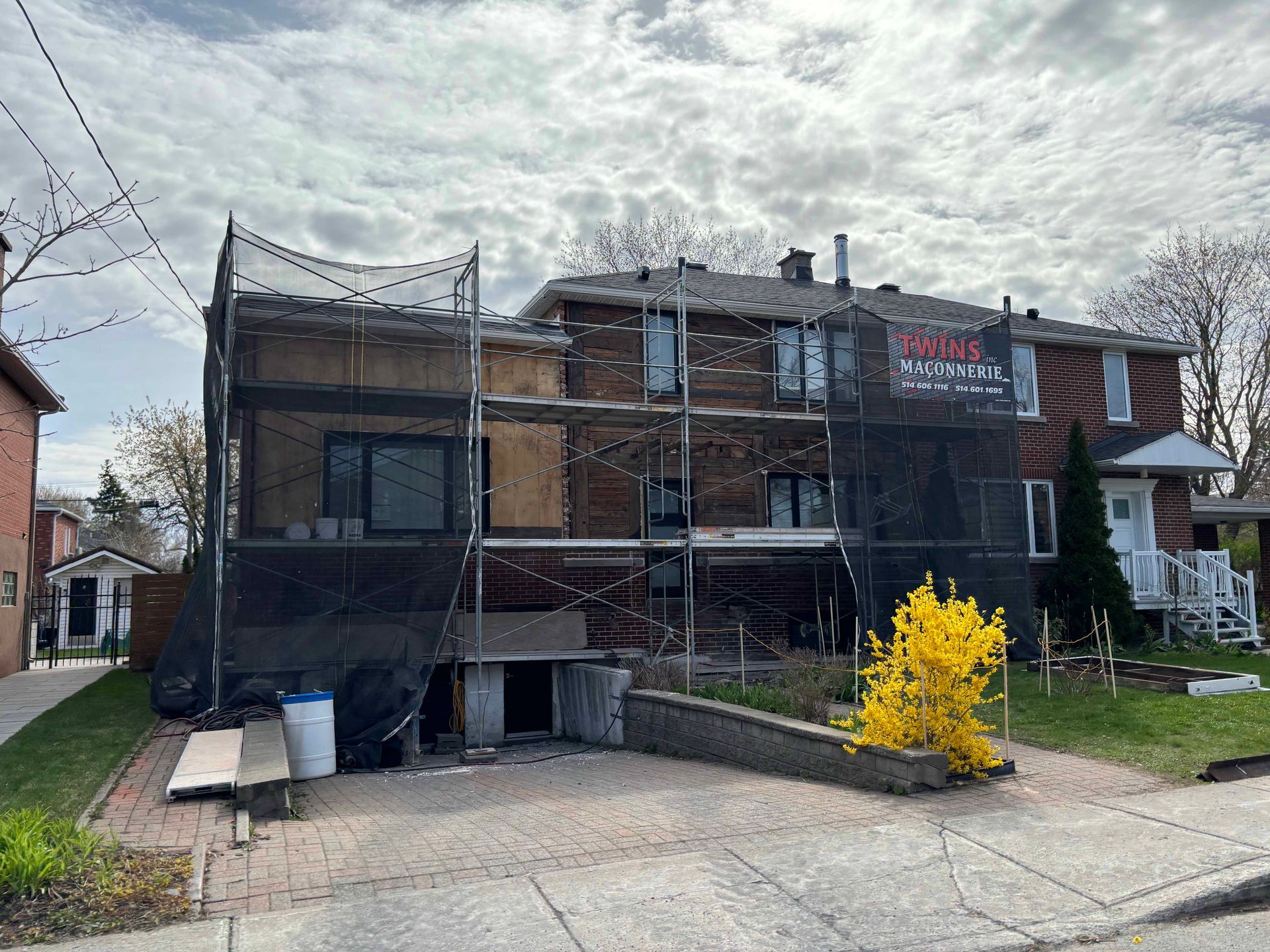 Bâtiment en rénovation, recouvert d'échafaudages et de filets. Bardage brun, ciel nuageux, buisson jaune.