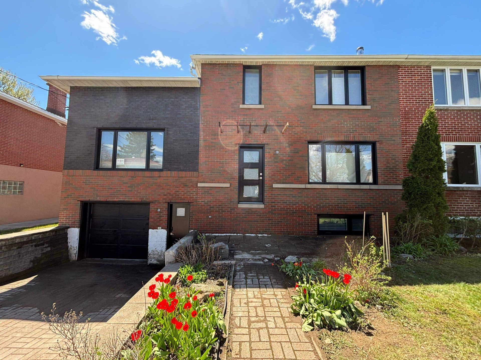 Bâtiment en briques avec porte de garage noire, allée en briques, tulipes rouges, ciel ensoleillé.