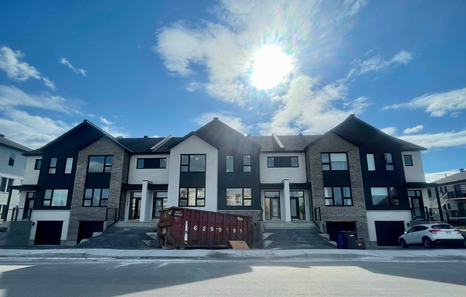 Rangée de maisons de ville neuves sous un soleil éclatant, bardage blanc et noir, toits sombres, débris de construction.