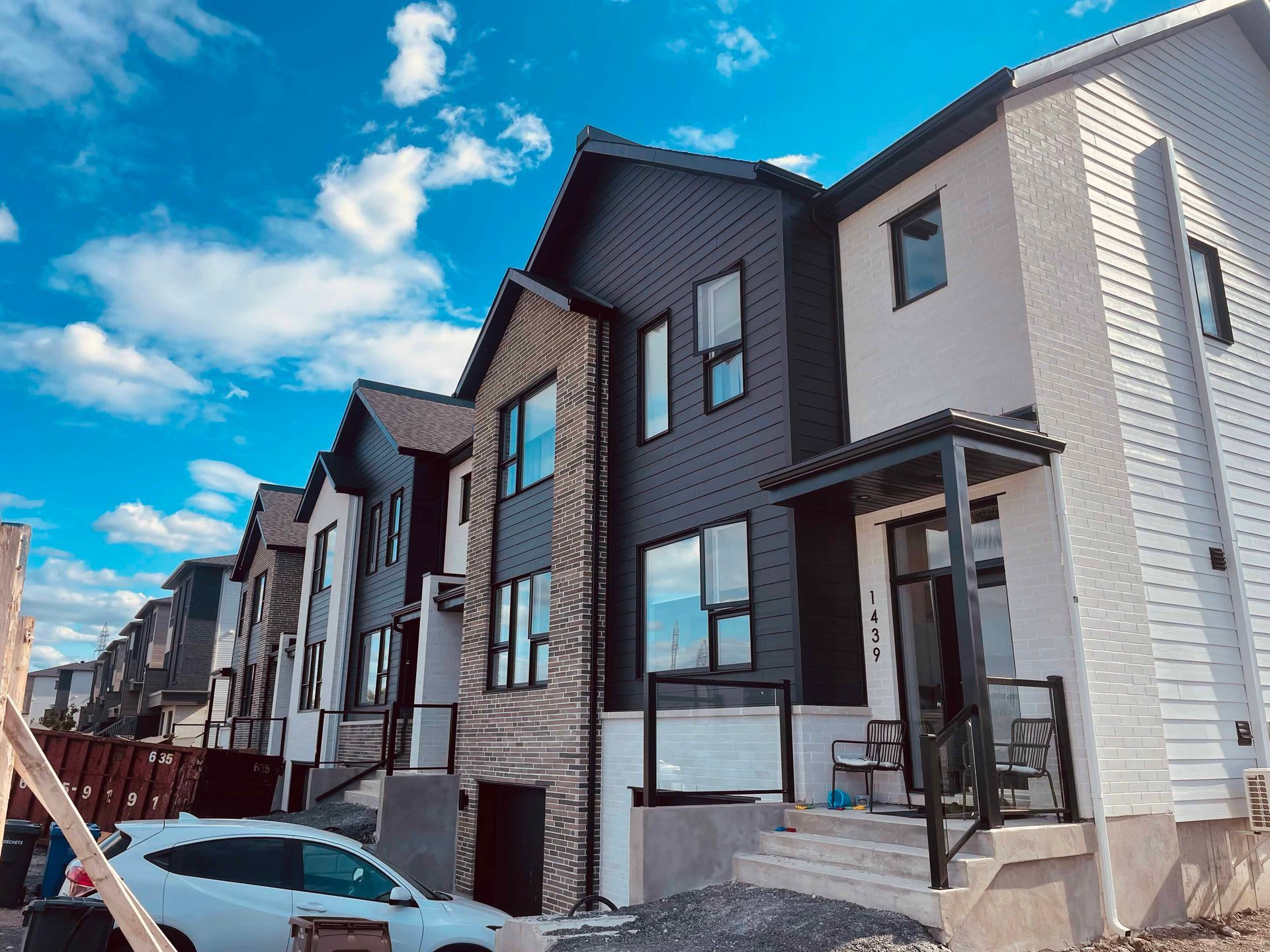 Maisons de ville aux façades sombres et aux grandes fenêtres, sous un ciel bleu éclatant parsemé de nuages.