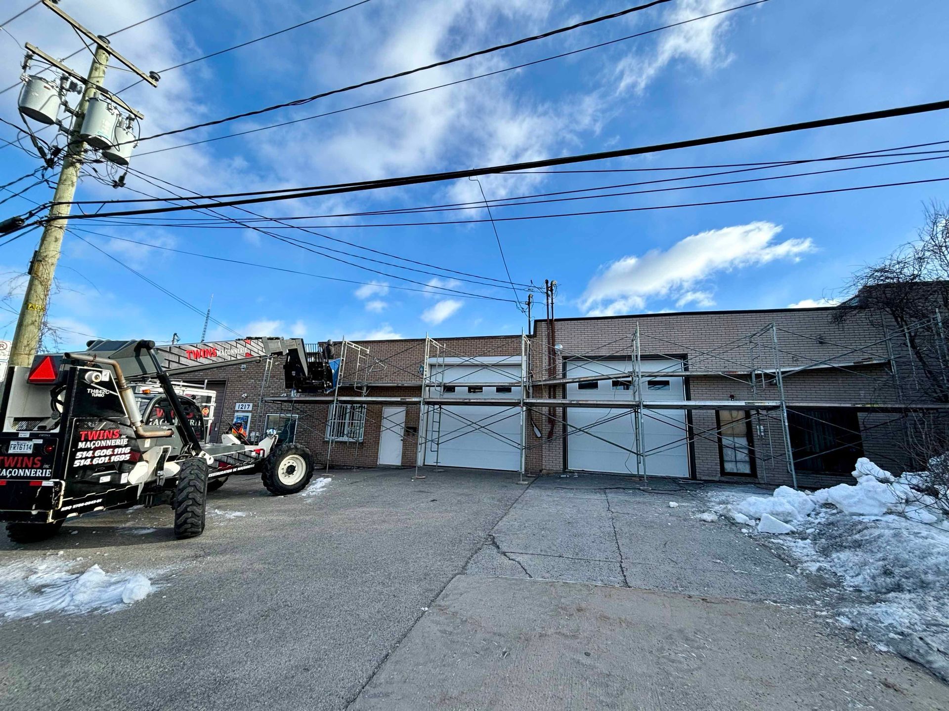 Vue extérieure d'un bâtiment avec portes de garage ouvertes et échafaudages, tracteur au premier plan, ciel nuageux.