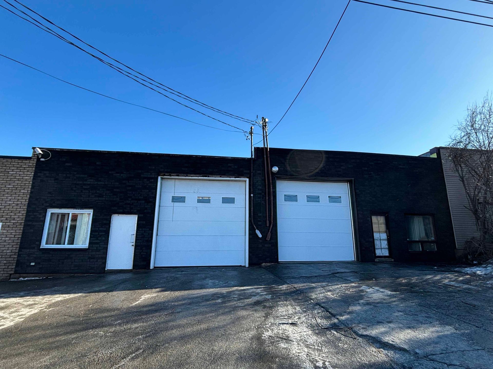 Bâtiment noir avec deux grandes portes de garage, une petite porte et une fenêtre se détachant sur un ciel bleu clair.